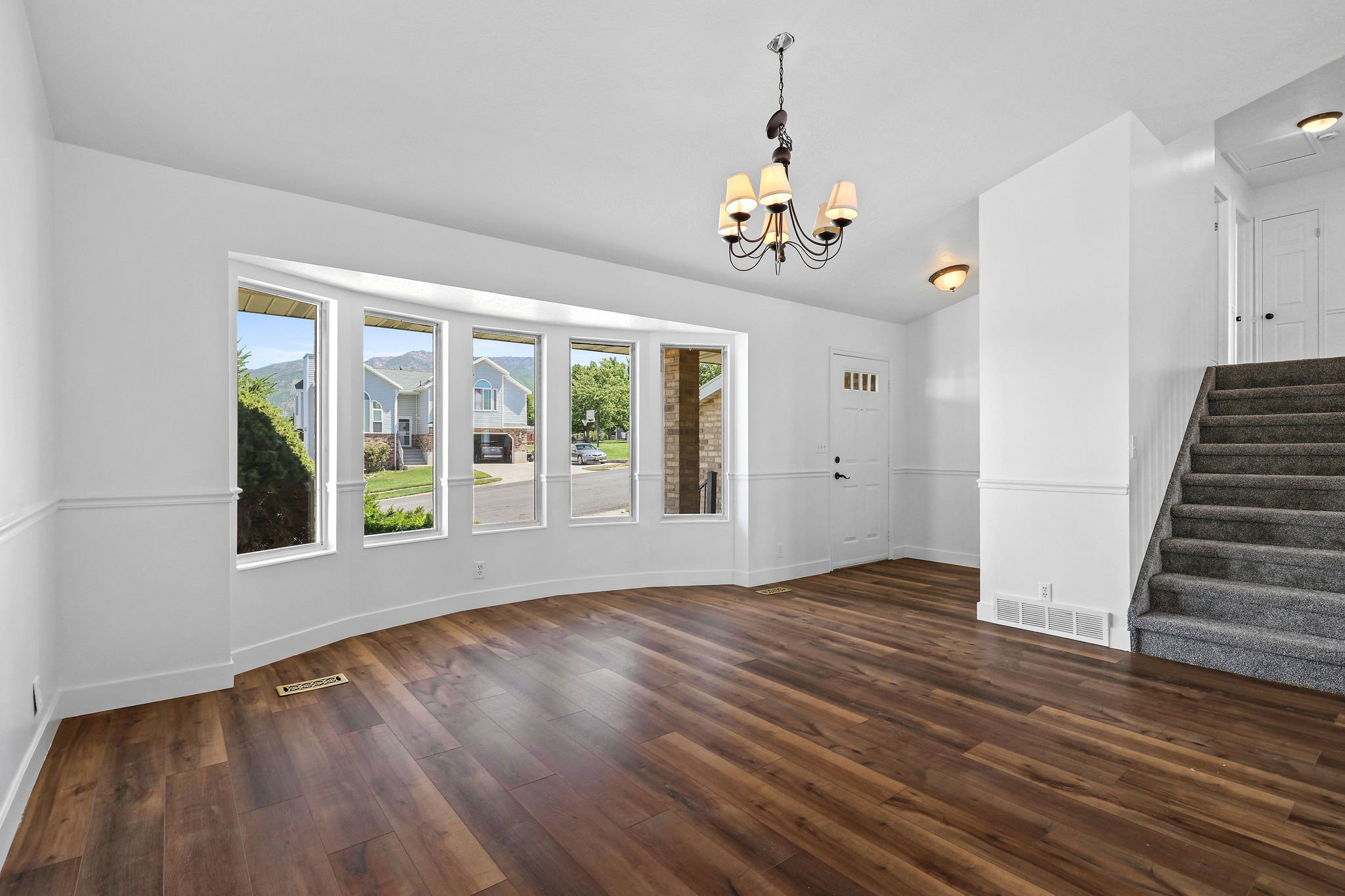 Open-concept living area featuring wood-finish flooring, a curved bay window, wainscoting, and a staircase with gray carpeting