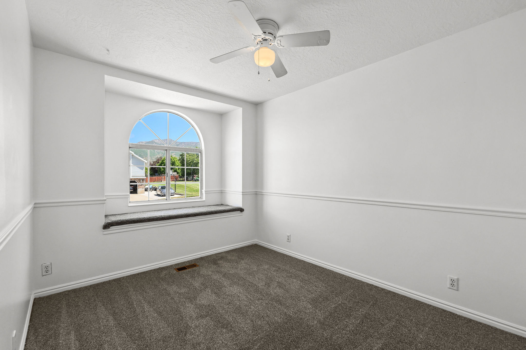 Arched window with integrated window seat, ceiling fan with light fixture, gray carpeting, and white baseboards