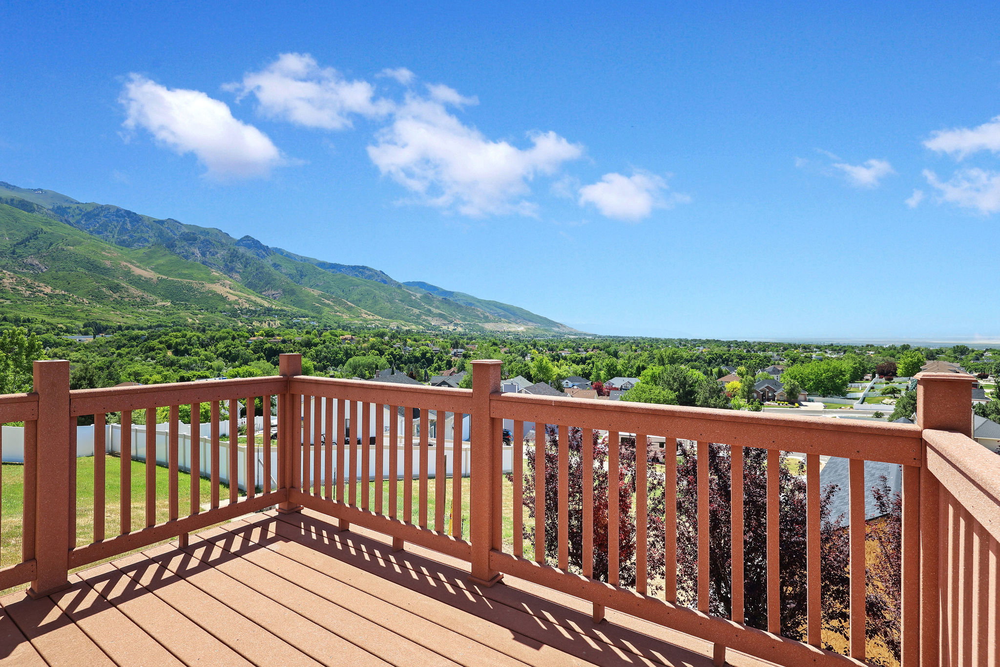 Outdoor deck with wood-finish railing and decking, featuring a panoramic landscape with distant mountains, green foliage, and a bright blue sky