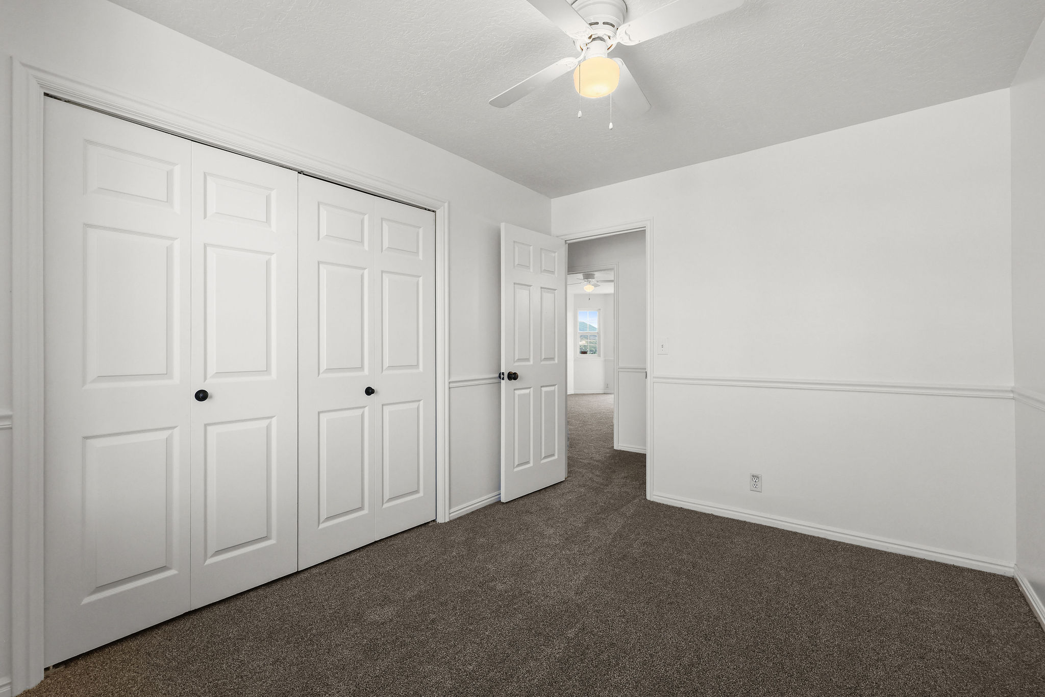 Bedroom featuring white paneled closet doors with black hardware, neutral wall-to-wall carpeting, white painted walls, and a ceiling fan with integrated lighting