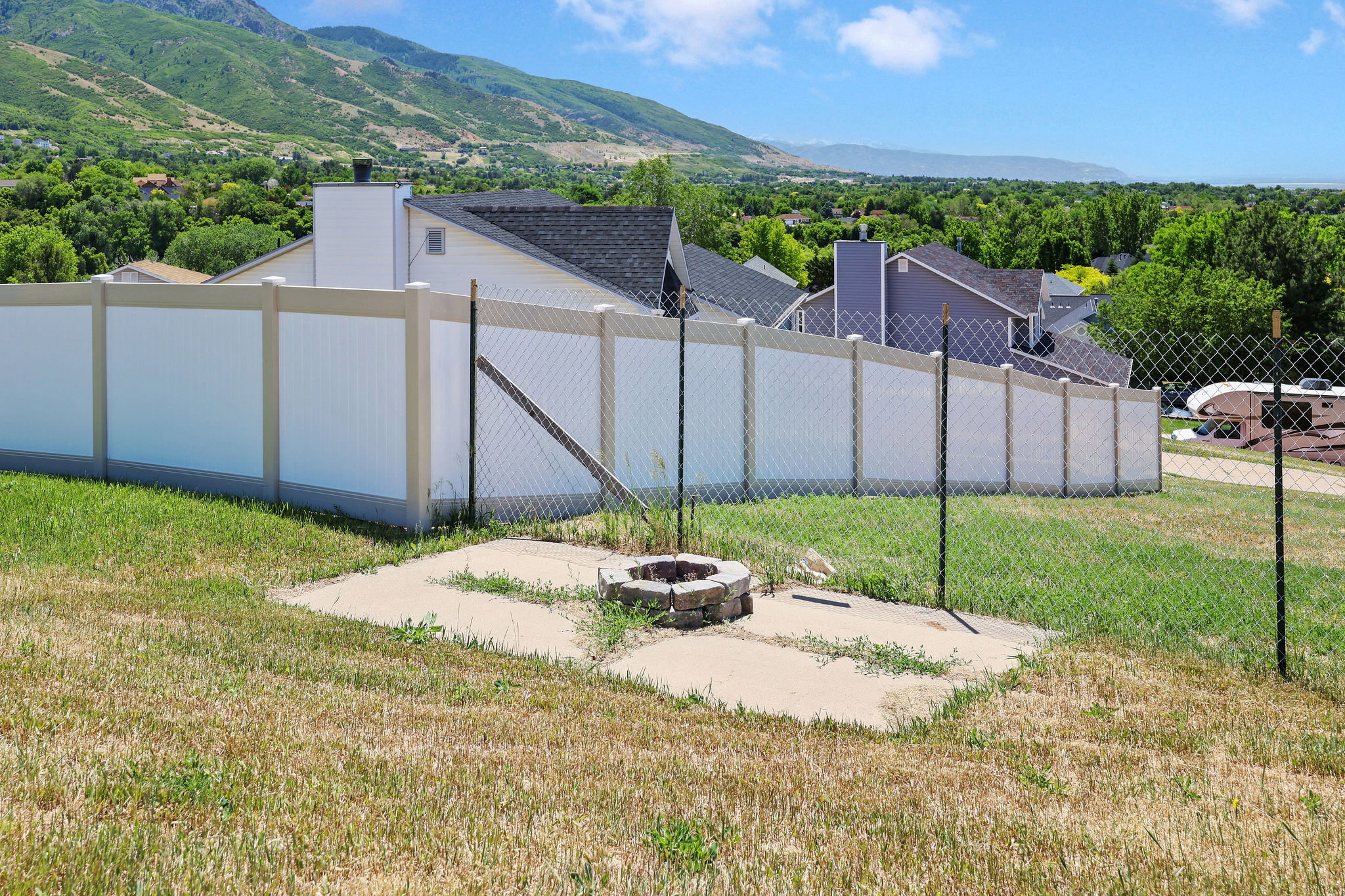 Hillside property featuring a stone-ring fire pit on a concrete pad, surrounded by a chain-link fence