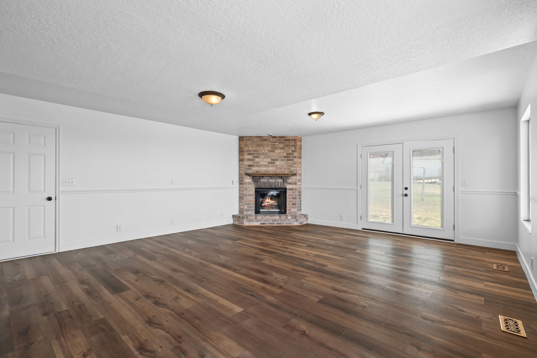Spacious room featuring wood-finish flooring, a brick-surround fireplace, dual French doors, a window, and wainscoting