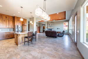 Expansive great room featuring a kitchen with custom wood cabinetry and a large island, an open living area with a stone-clad fireplace, tile flooring, and multiple large windows