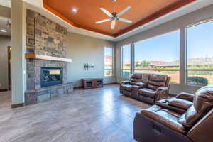 Spacious living area featuring a stone-clad fireplace with a wood mantel, large windows, a tray ceiling with a ceiling fan, recessed lighting, and tile flooring