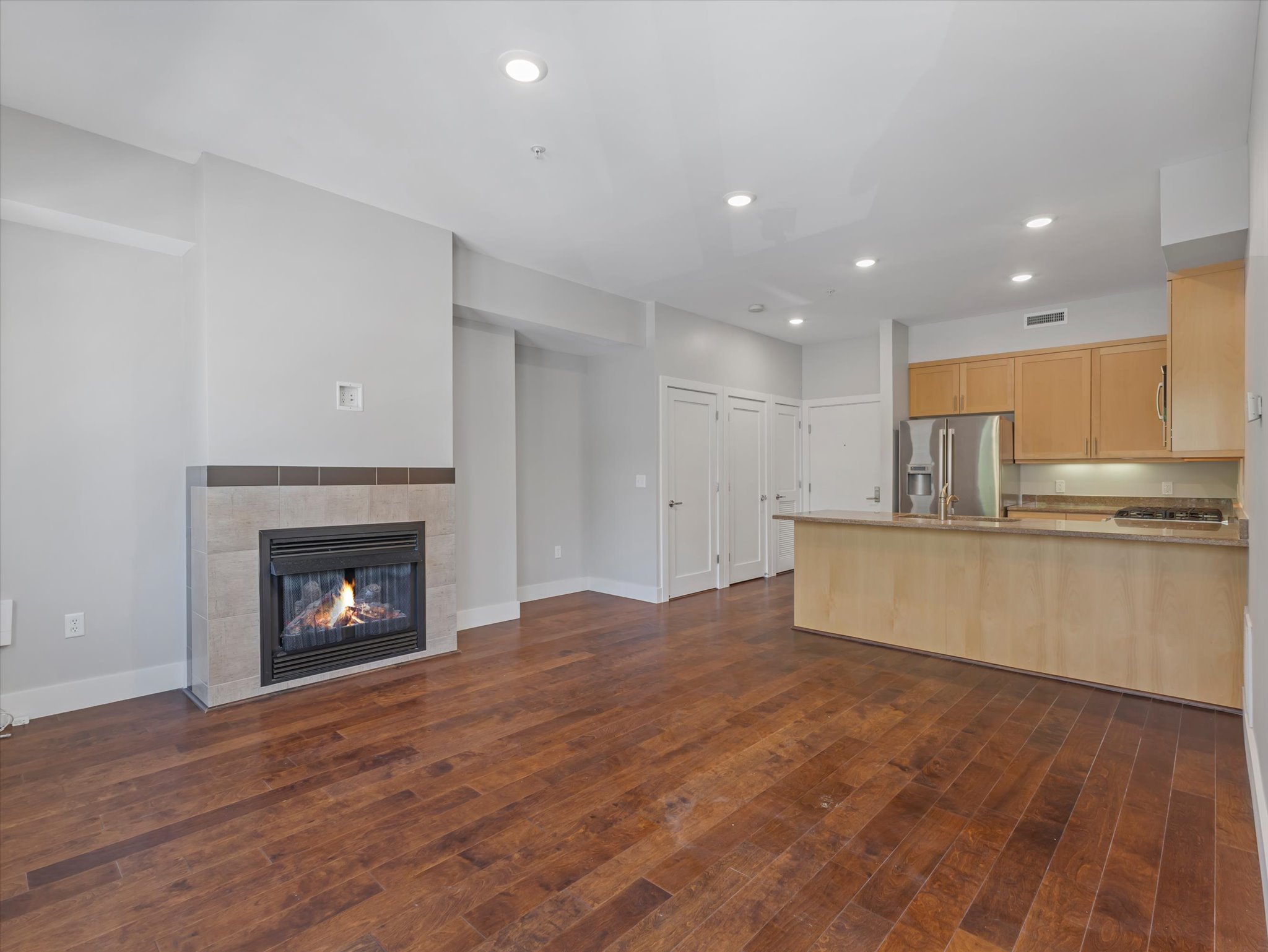 Open-concept living area featuring wood-finish flooring, a tiled fireplace, recessed lighting, and a kitchen with warm-toned cabinetry, stainless steel appliances, and a breakfast bar