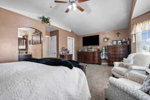 Carpeted room featuring a vaulted ceiling with a ceiling fan, a window with louvered blinds, and an arched doorway leading to a vanity area with a mirror