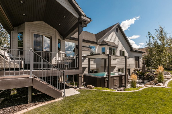 Expansive backyard featuring a manicured lawn, a multi-level deck with metal railings, a built-in hot tub with an overhead pergola, and a stone-lined garden bed with mature landscaping