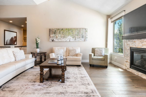 Living area featuring vaulted ceilings, light wood-finish flooring, a stacked stone fireplace with a dark wood mantel, and large windows providing natural illumination