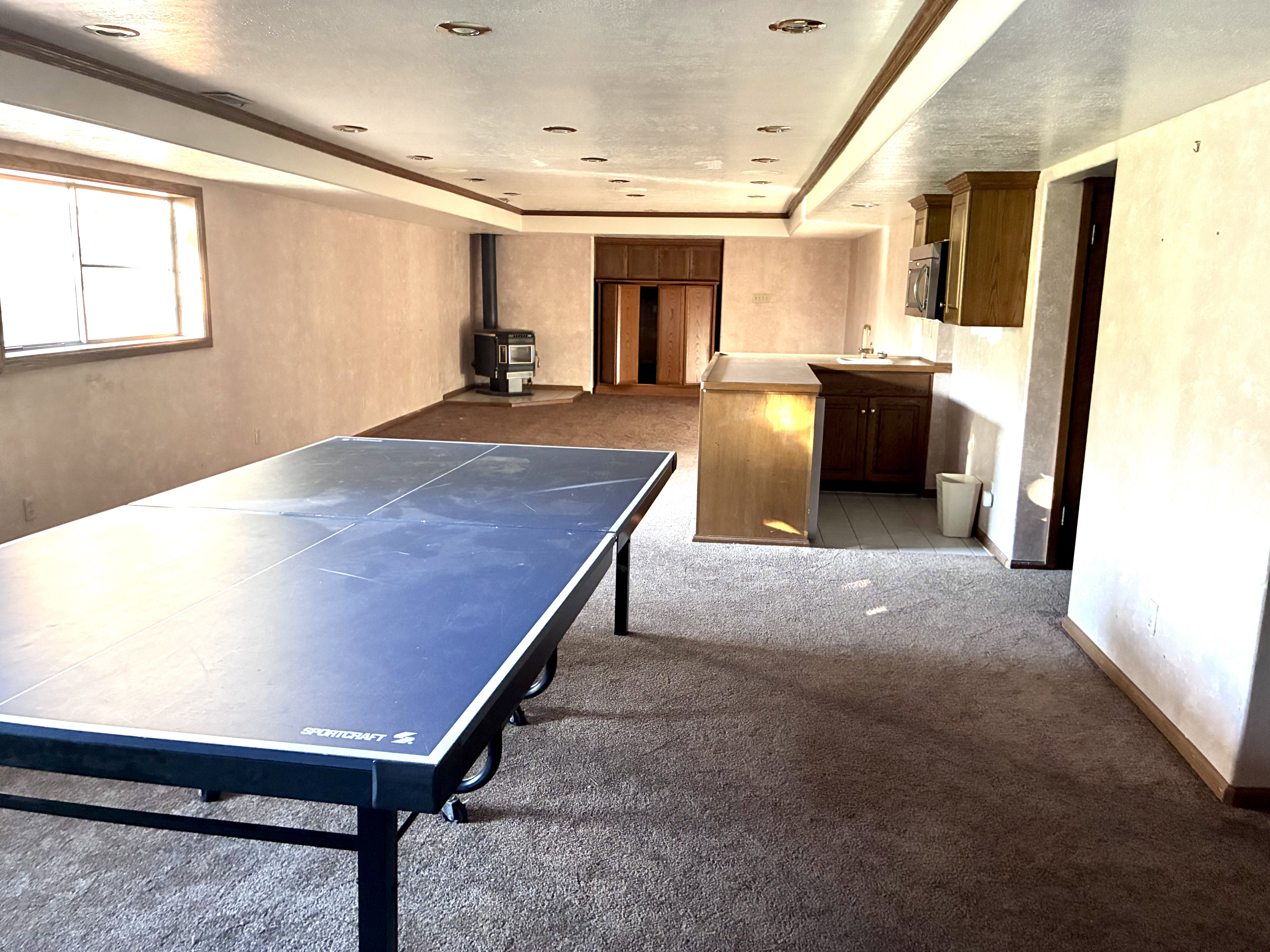 Spacious recreation room featuring recessed lighting, a built-in wet bar with wood cabinetry, a wood-burning stove, and ample natural light from a large window