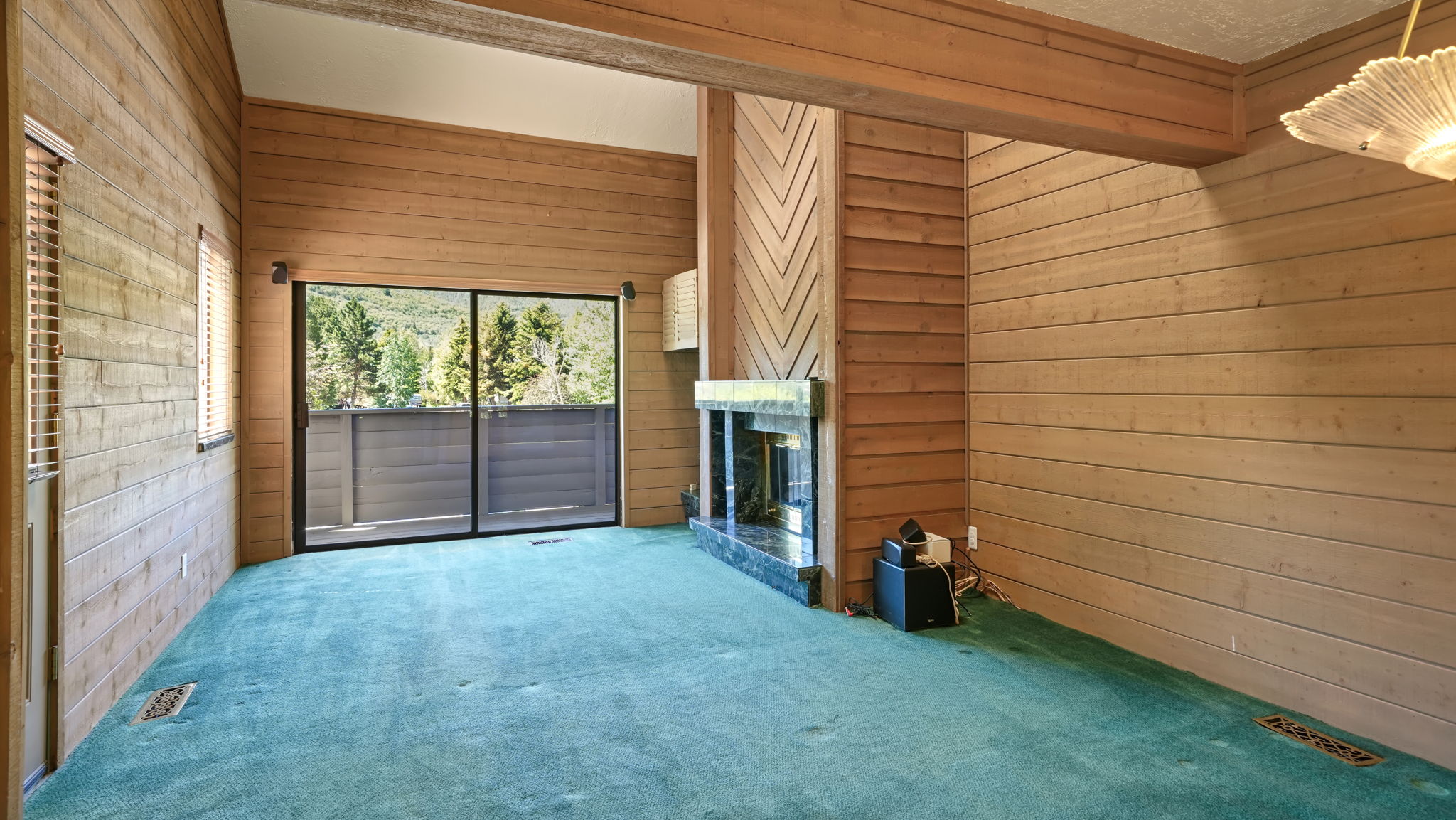 Spacious room with extensive wood-paneling, a large fireplace featuring a stone surround, and floor-to-ceiling sliding glass doors opening to an exterior deck