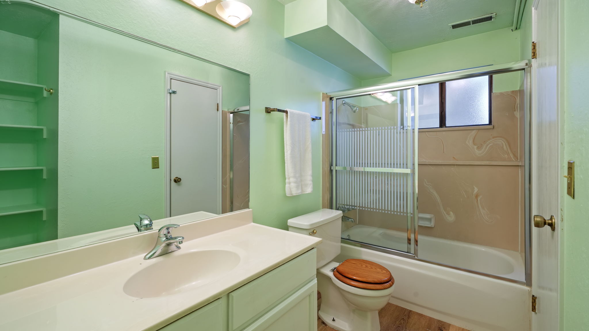 Bathroom with a light green color palette featuring a vanity with an integrated sink and chrome faucet, a large wall-mounted mirror, built-in shelving, and a tub/shower combination with a sliding glass enclosure and window
