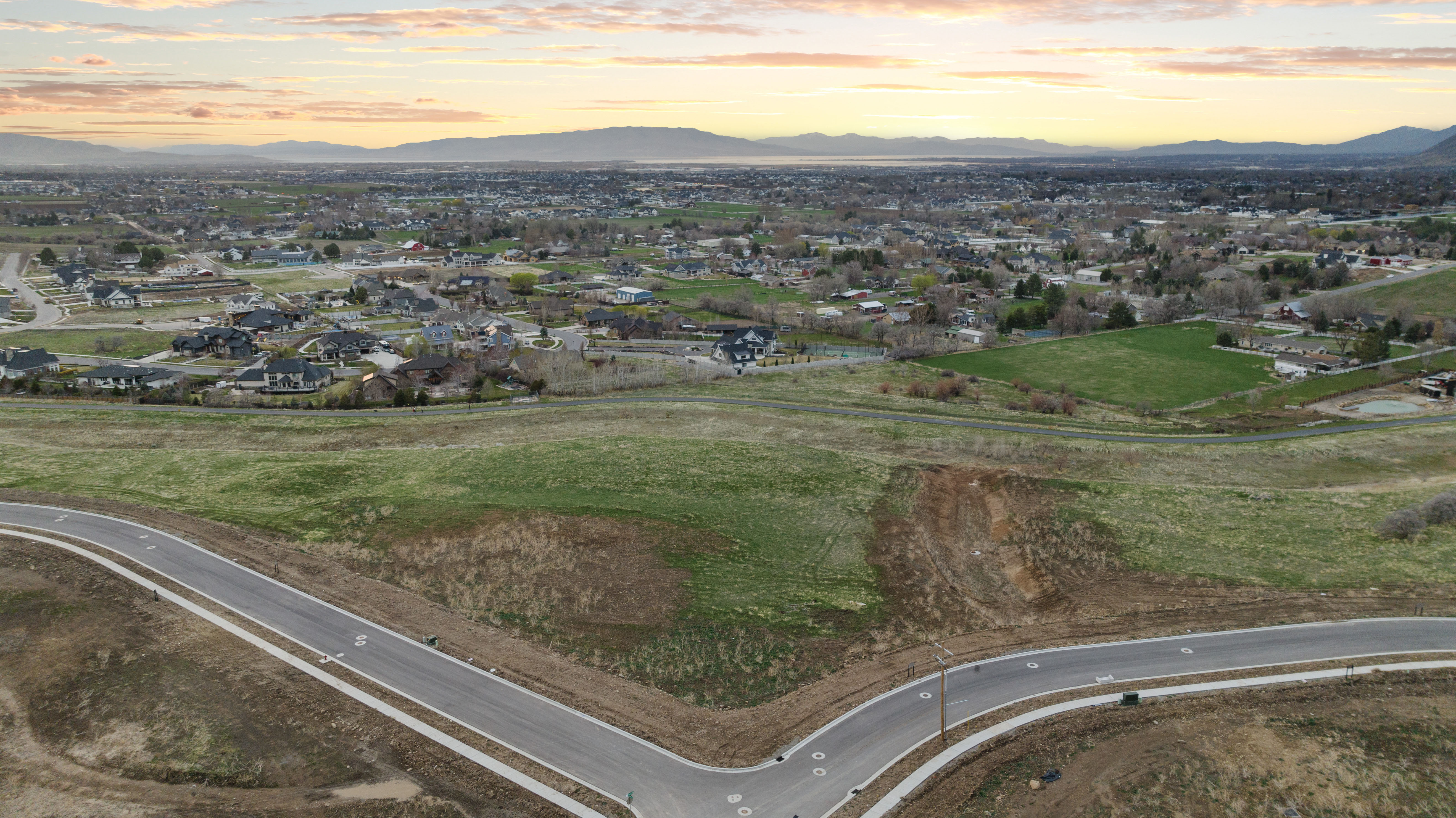 Expansive undeveloped land featuring a paved road with concrete sidewalks, established infrastructure, and distant residential areas
