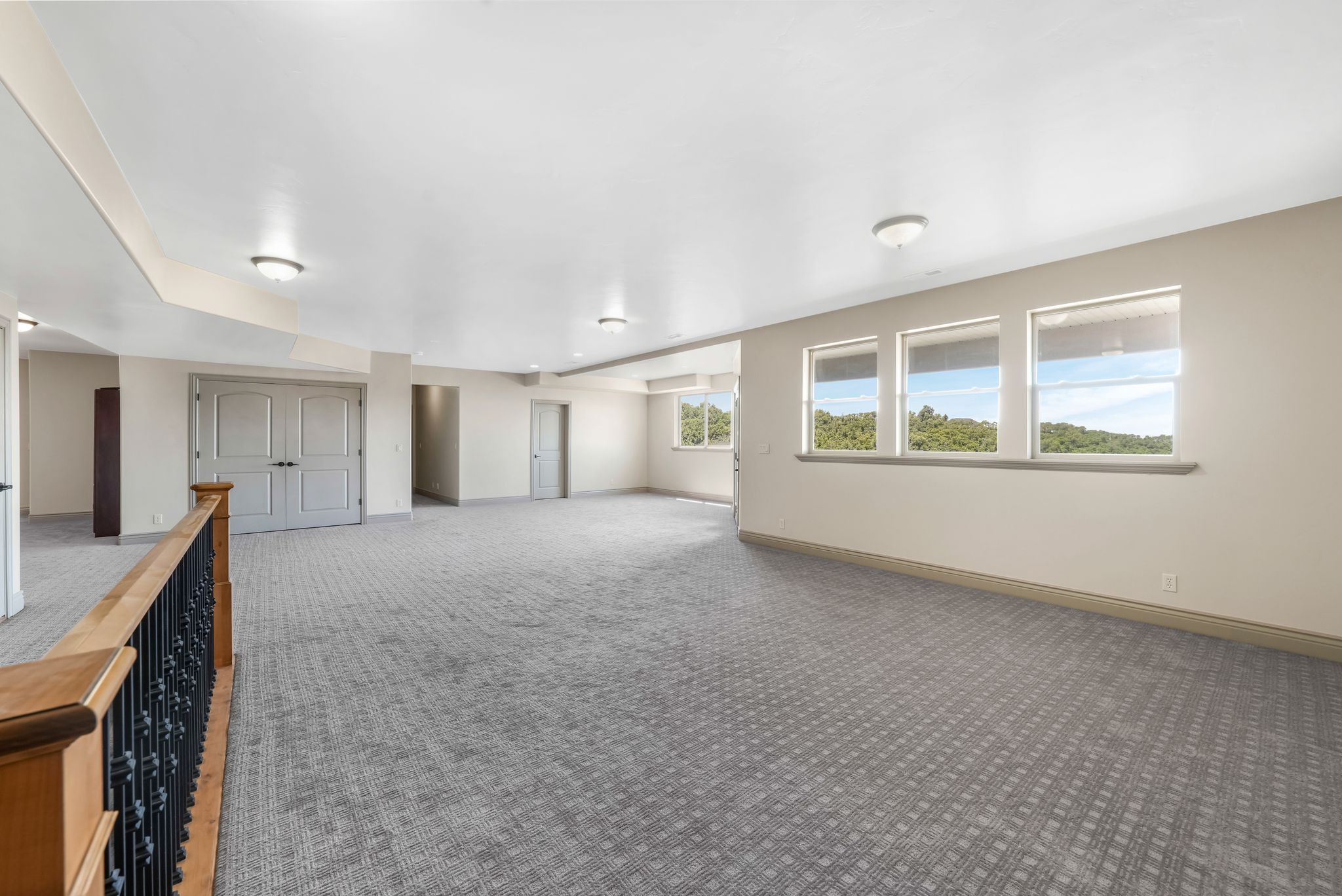 Expansive room featuring patterned carpeting, multiple windows with a scenic backdrop, light-toned walls, recessed lighting, and a hardwood banister with wrought iron spindles