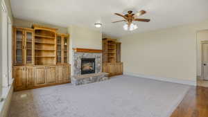 Living area featuring a stone fireplace, built-in wooden cabinetry with glass-front doors, a ceiling fan with integrated lighting, and carpeted flooring transitioning to wood-finish flooring