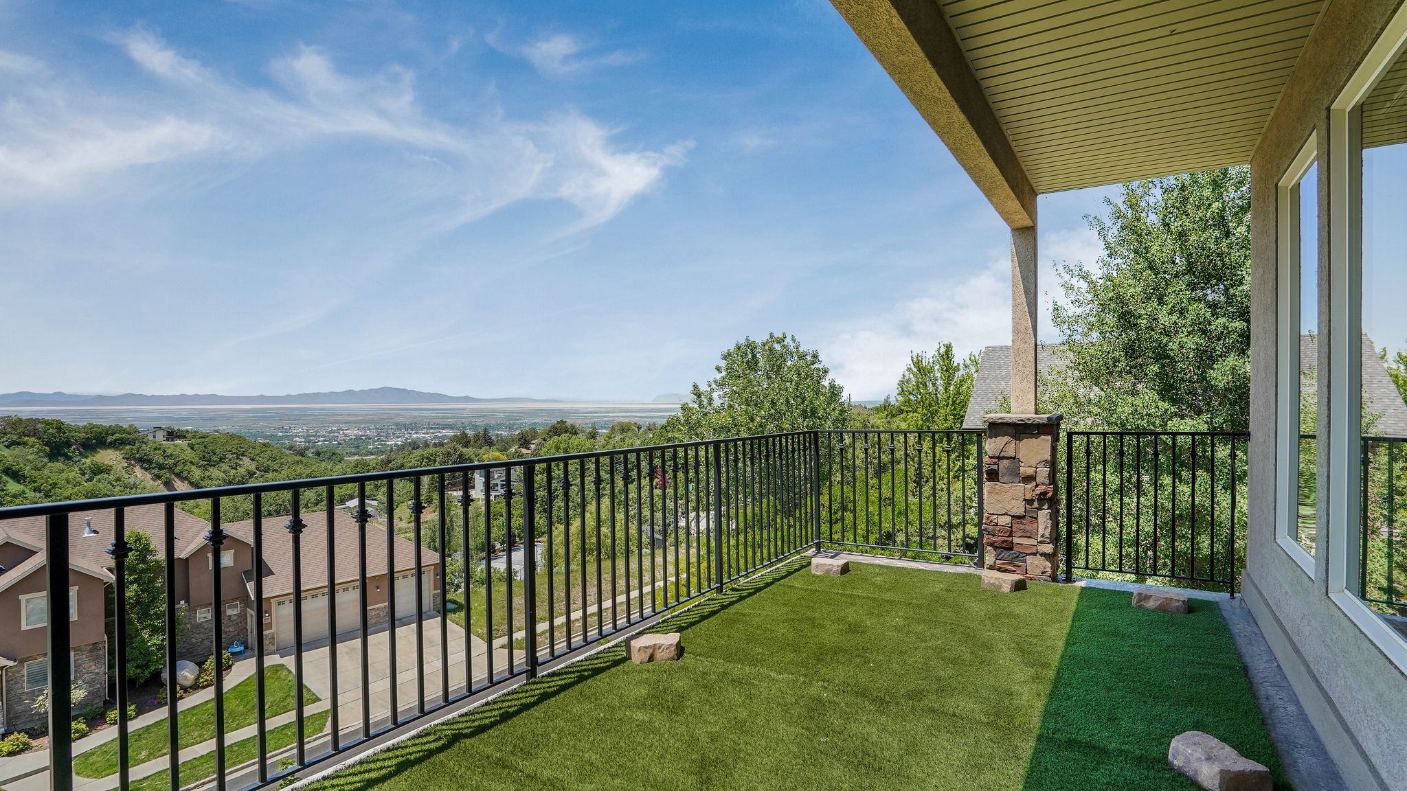 Covered balcony featuring artificial turf flooring, black metal railings, a stone-clad column, and large windows