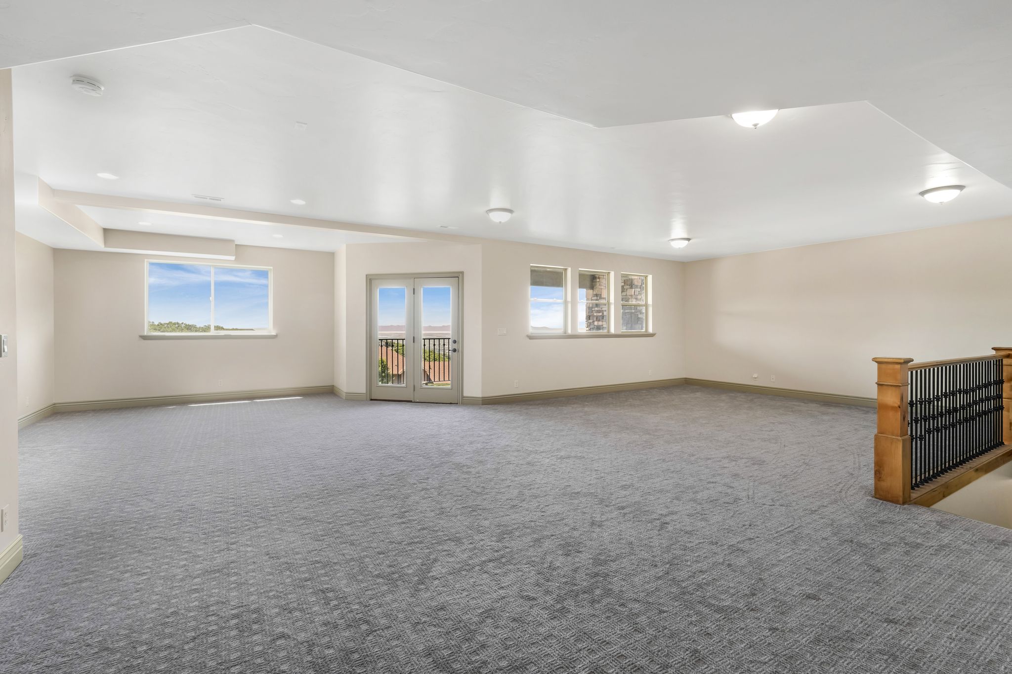 Expansive carpeted room featuring multiple windows, French doors with a balcony, and a wooden staircase with black iron balusters