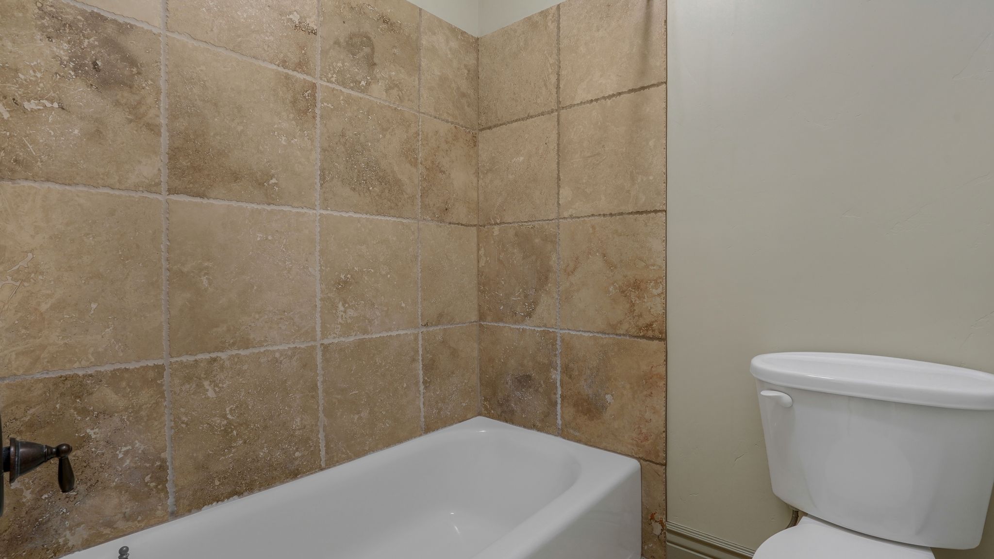 Full bathtub with a bronze-finish fixture, surrounded by large format beige wall tiles and neutral painted walls