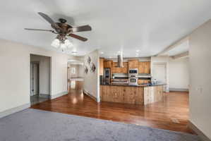 Open-concept kitchen featuring wood cabinetry, stainless steel appliances, dark countertops, and a wood-paneled island with a breakfast bar
