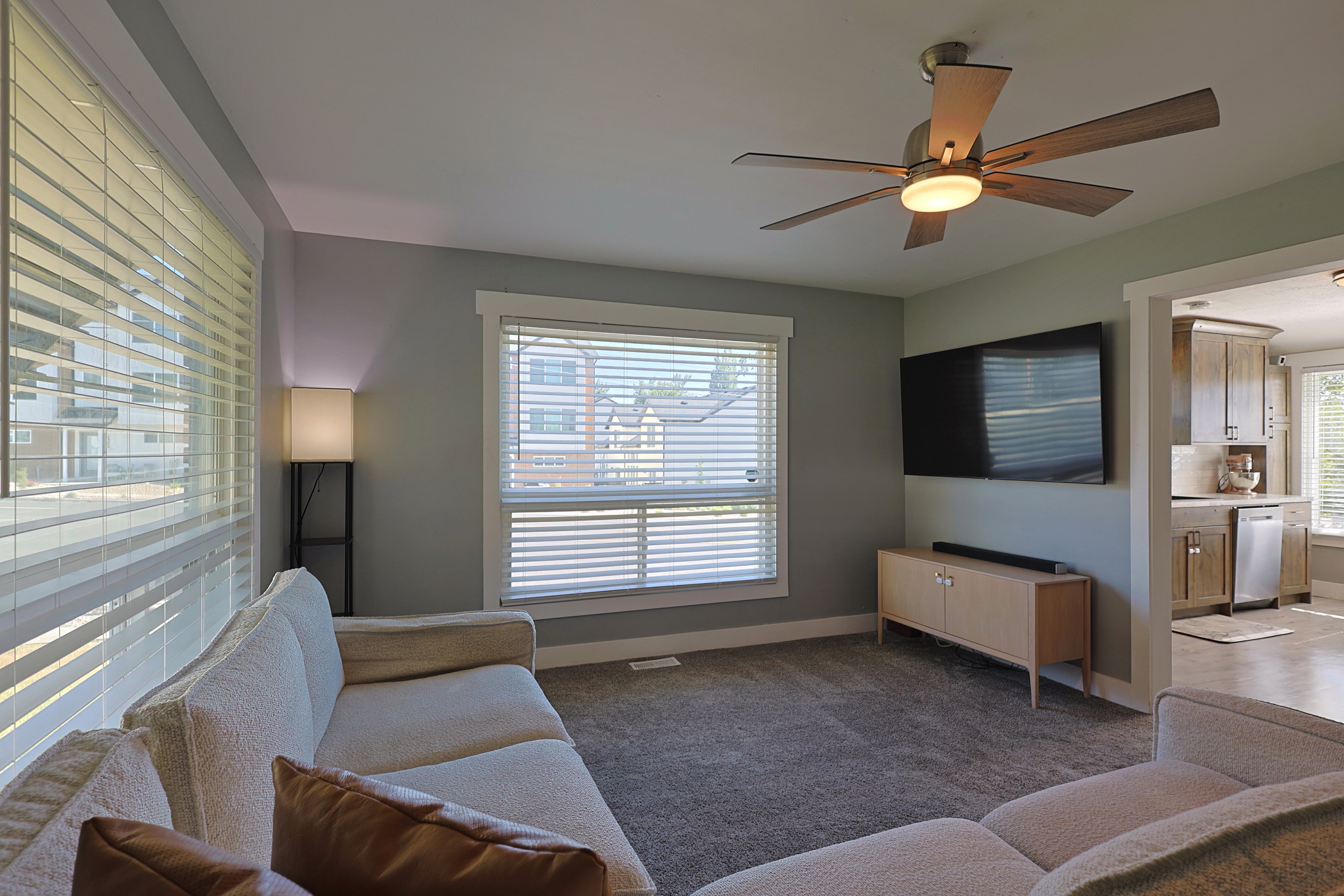 Carpeted living area featuring multiple windows with blinds, a ceiling fan with integrated lighting, and a wide entryway leading to a kitchen with wood-finish cabinetry
