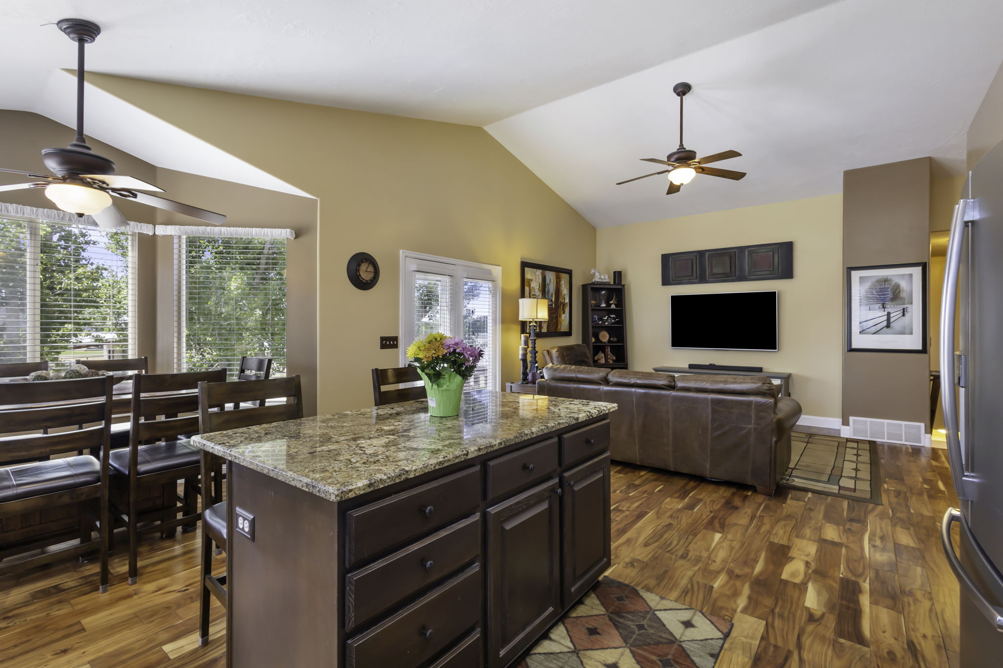 Open-concept living space with vaulted ceilings, cooling ceiling fans, hardwood  flooring, and a kitchen island featuring granite countertops.