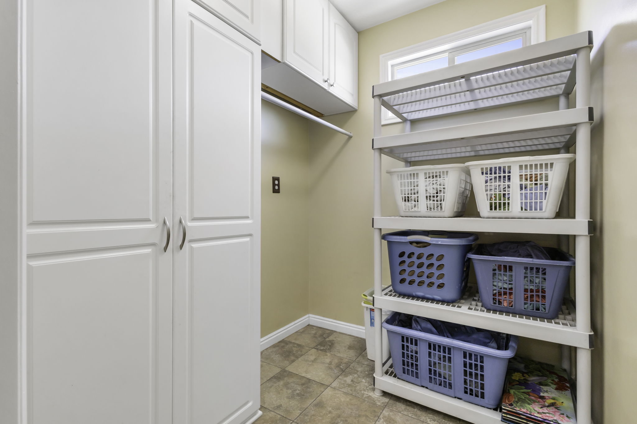 Spacious laundry area featuring white storage cabinetry, a built-in hanging rod, a window for natural light, and tile flooring