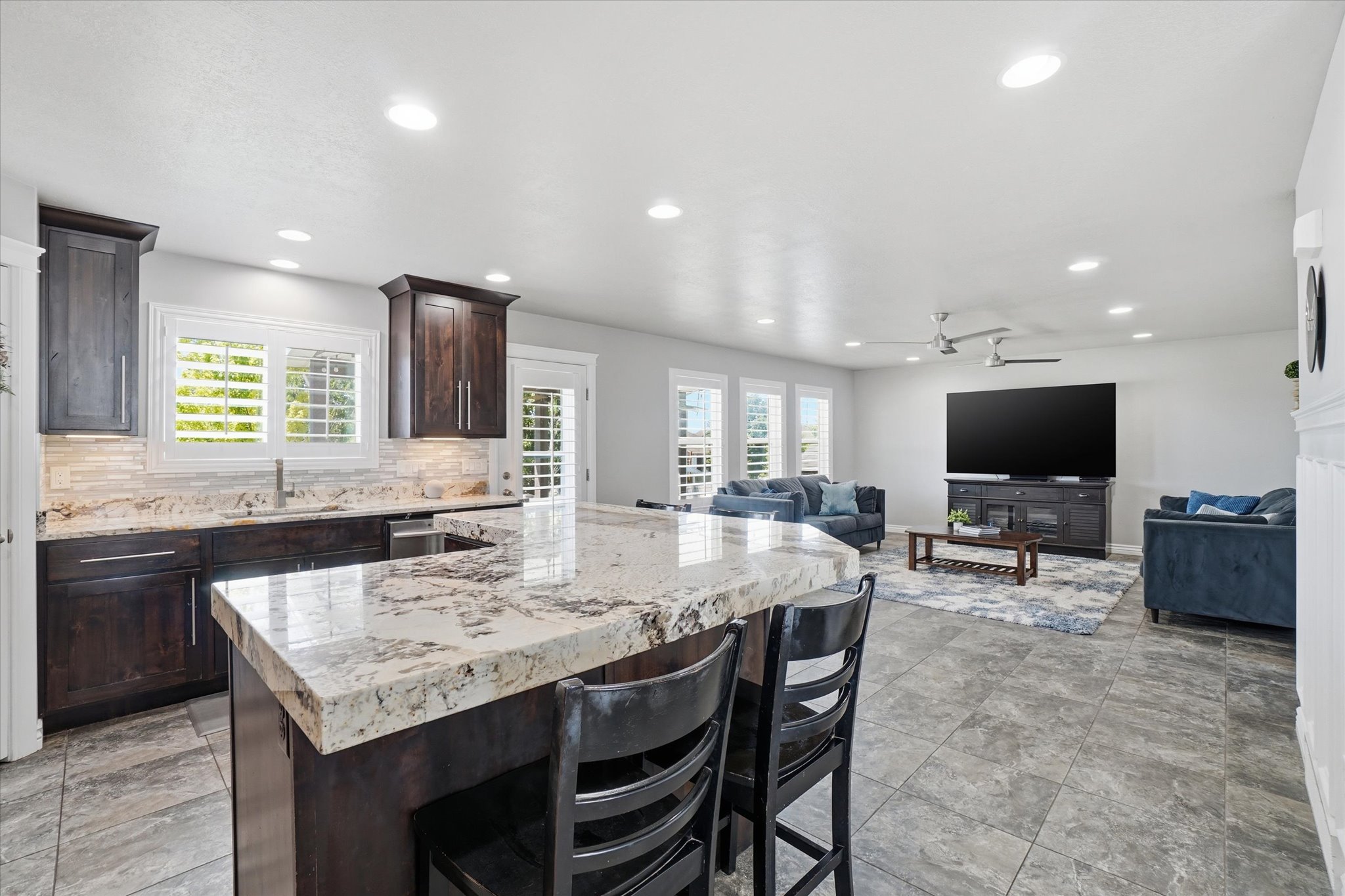 Open-concept kitchen and living area featuring granite countertops, dark wood cabinetry, white plantation shutters, tiled flooring, and recessed lighting