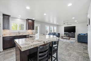 Open-concept kitchen and living area featuring granite countertops, dark wood cabinetry, white plantation shutters, tiled flooring, and recessed lighting