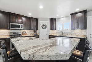 Spacious kitchen featuring a large island with a substantial countertop, dark wood-finish cabinetry, stainless steel appliances, a tile backsplash, and recessed lighting