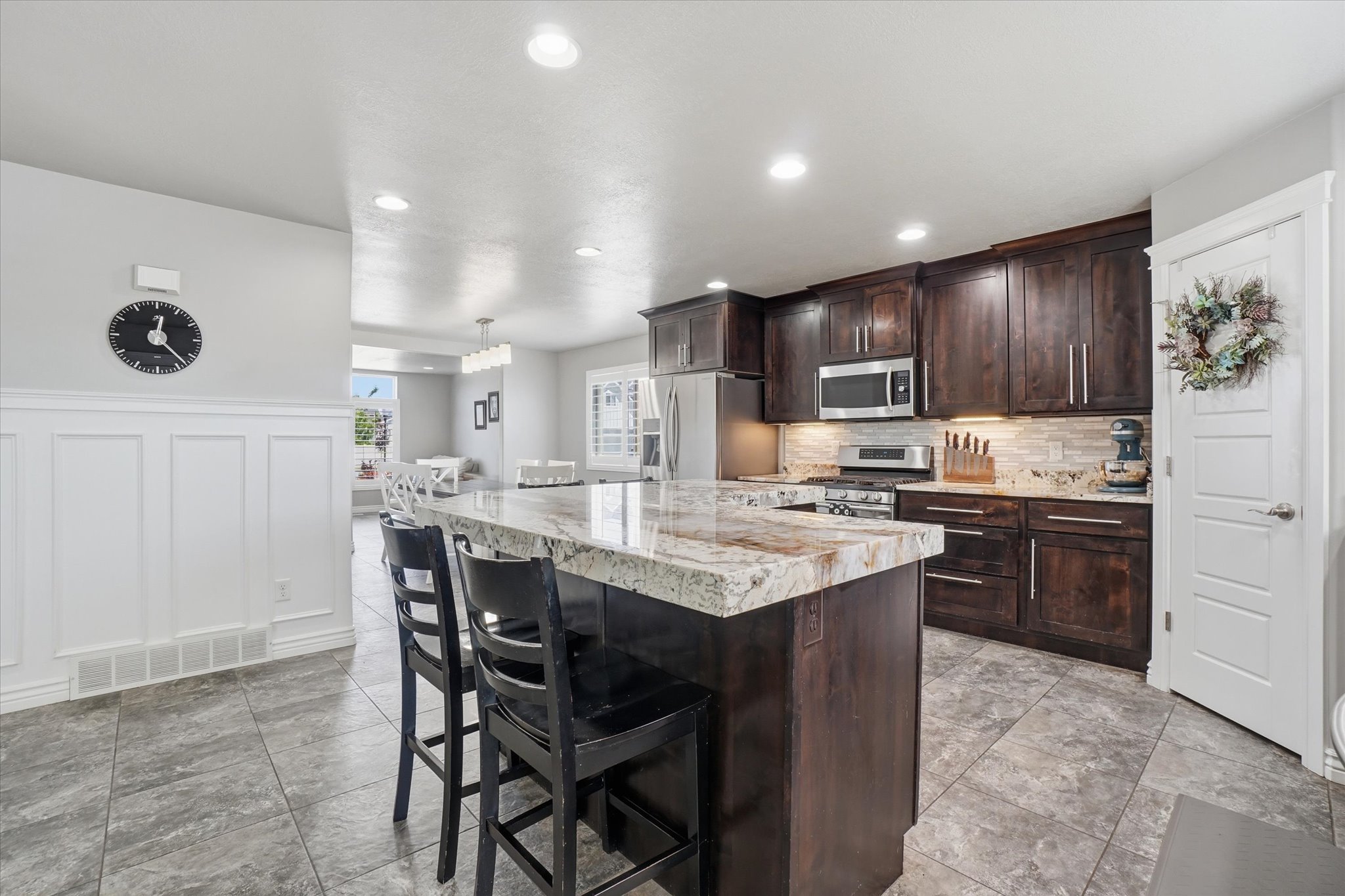Open-concept kitchen featuring dark wood cabinetry, stainless steel appliances, a multi-tone stone backsplash, and a central island with a light-colored stone countertop