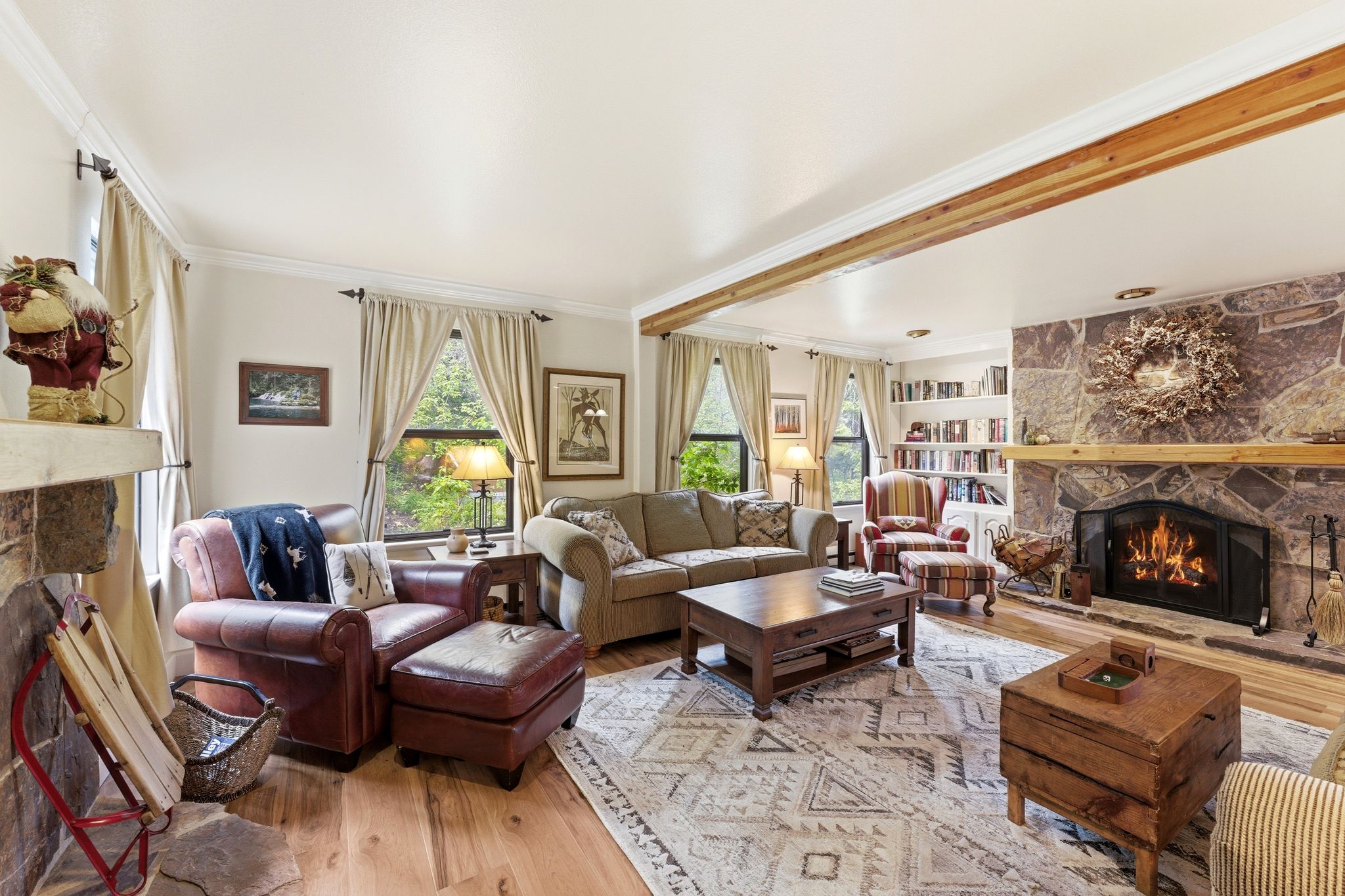 Spacious living area featuring a stone-clad fireplace with a natural wood mantle, light wood-finish flooring, and exposed ceiling beams