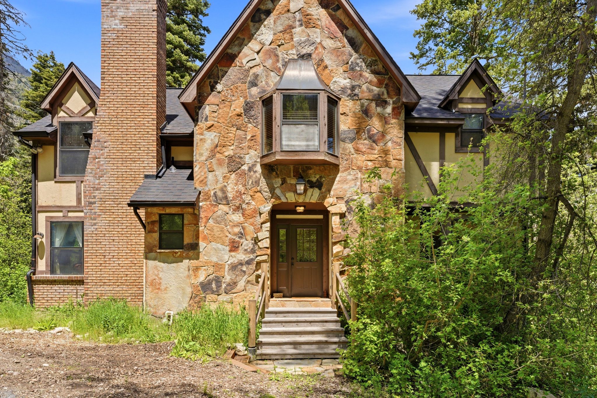 Stone and stucco Tudor-style facade featuring a prominent stone chimney, a gabled roofline, double entry doors, and mature surrounding foliage