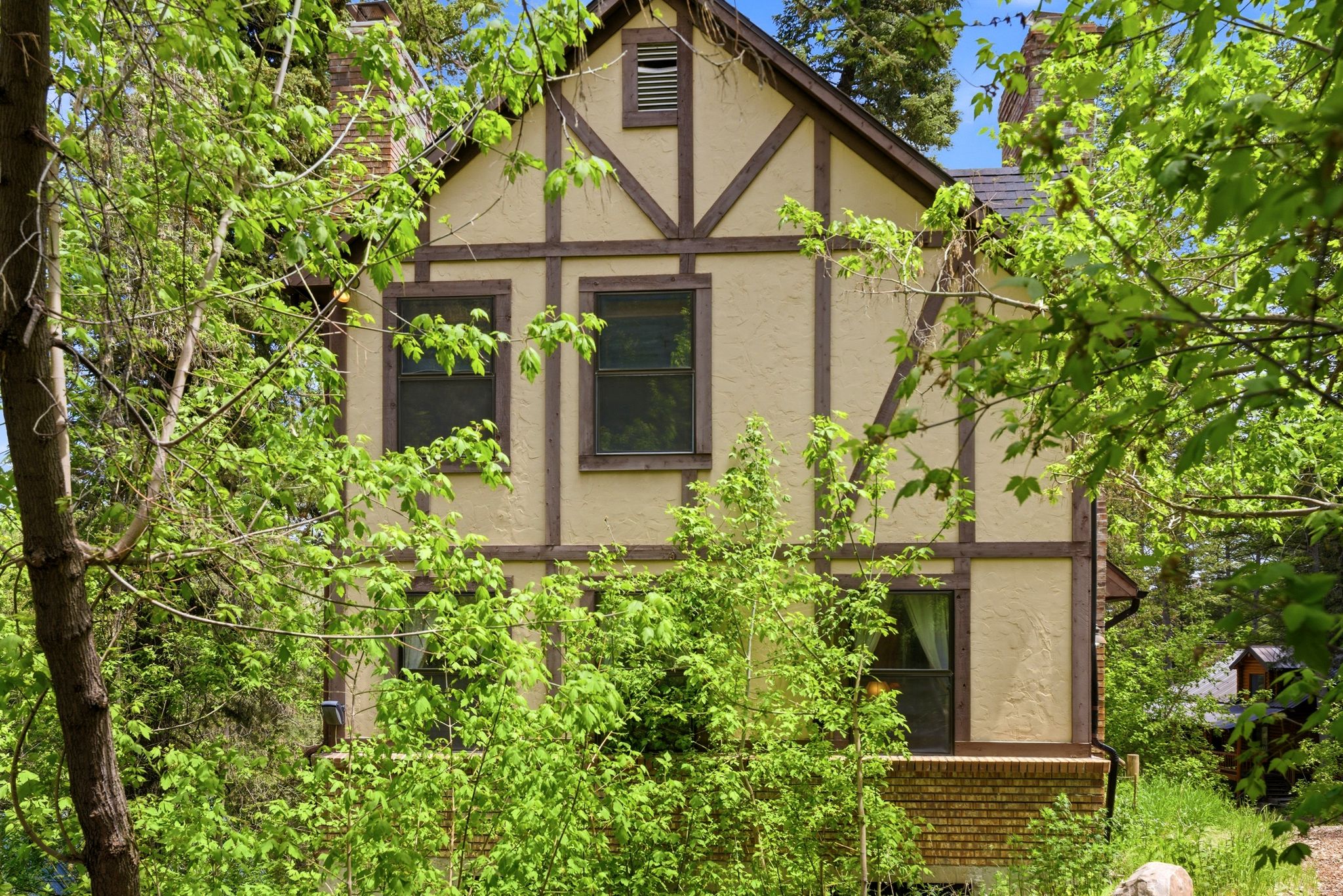Tudor-style exterior featuring cream stucco with dark wood trim, multiple windows, and a visible portion of a brick foundation
