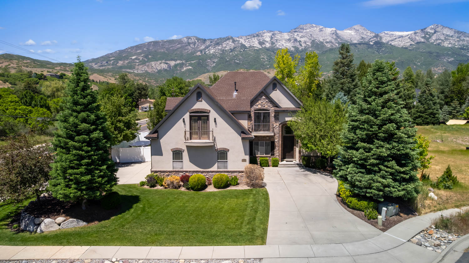 Stucco and stone exterior home featuring a curved driveway, established landscaping, and a mountainous backdrop