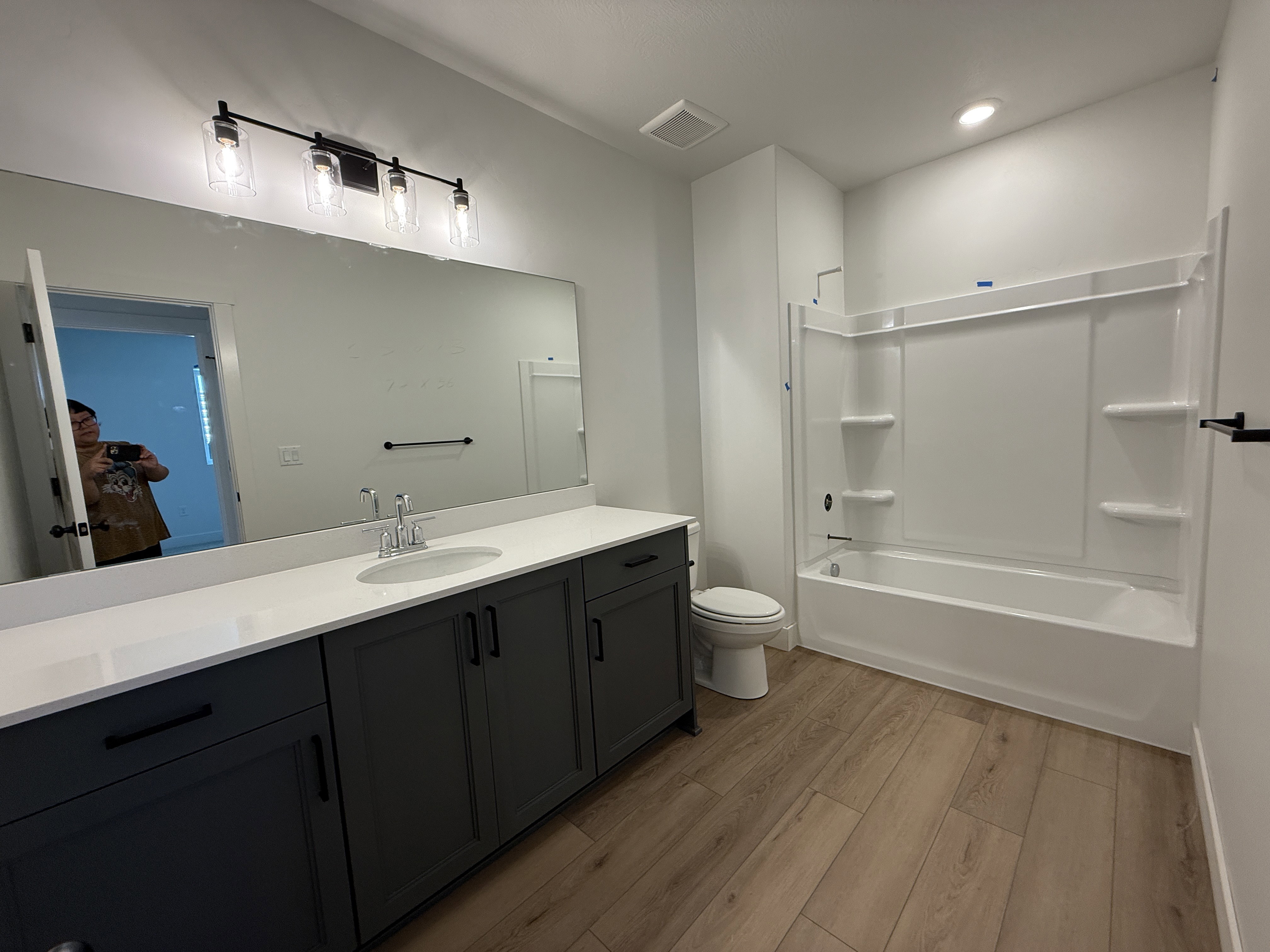 Full bathroom featuring a single-sink vanity with dark cabinetry and a light-toned countertop, a full-size bathtub with an integrated shower, wood-finish flooring, and a modern four-light vanity fixture