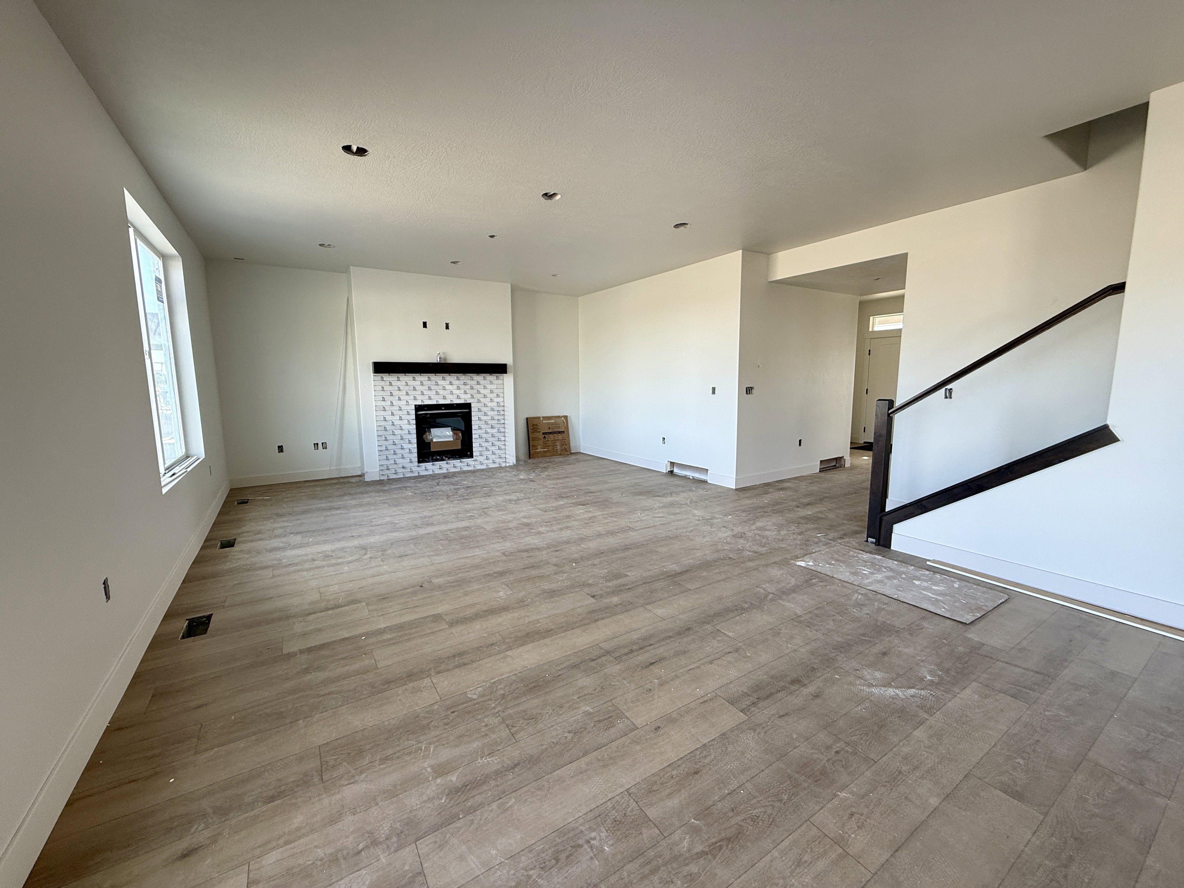 Spacious living area featuring wood-finish flooring, a white tiled fireplace with a black mantel, a large window, and a dark-stained staircase