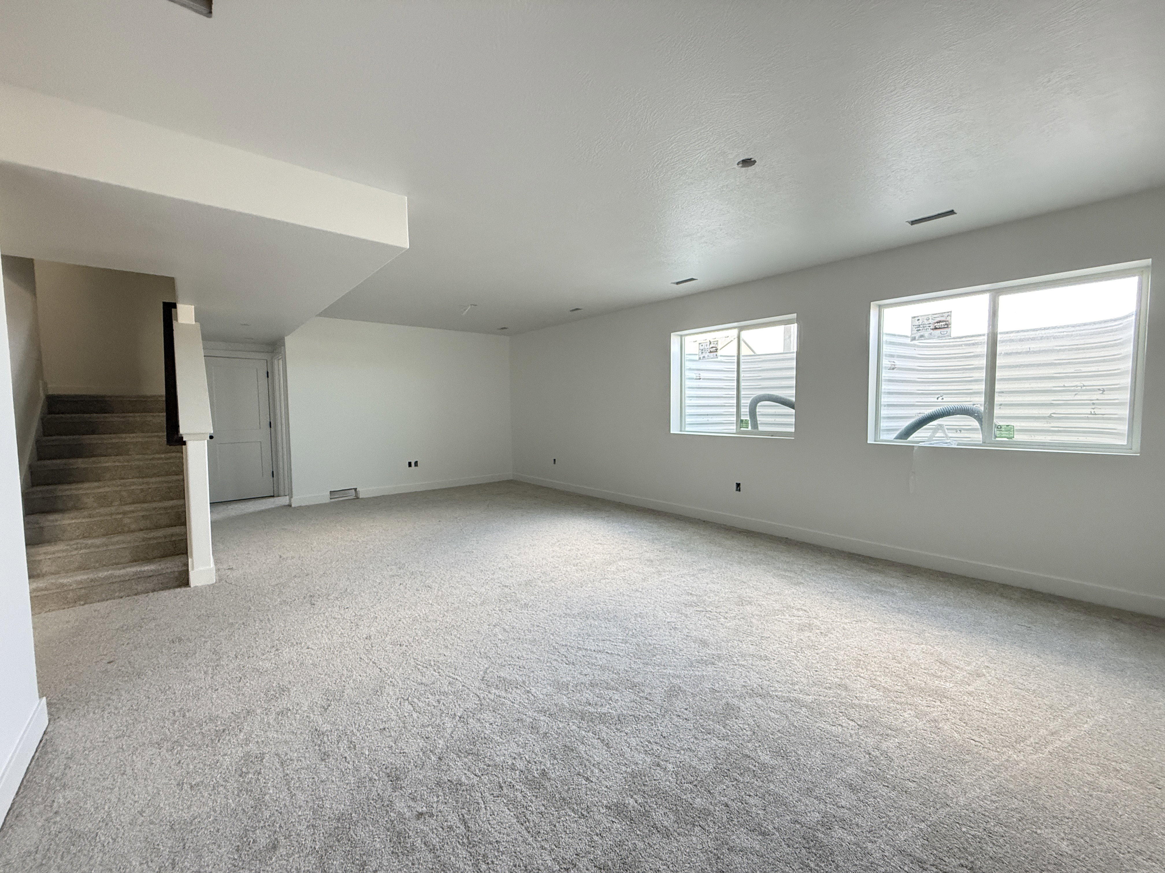 Spacious room featuring new carpet, white walls, two egress windows, a door, and a carpeted staircase with a white railing