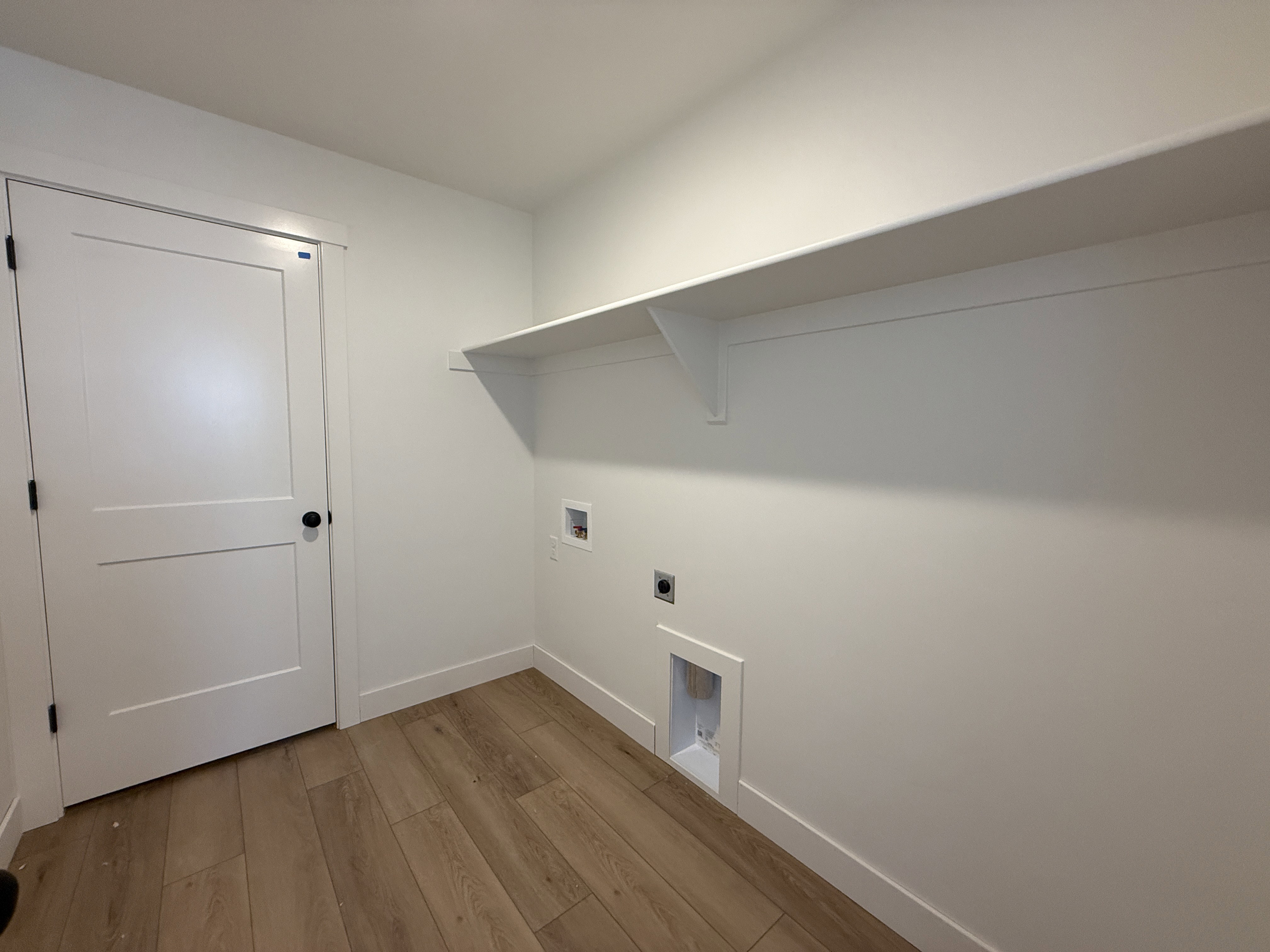 Dedicated utility space featuring wood-finish flooring, a contemporary white door with black hardware, and an integrated wall shelf