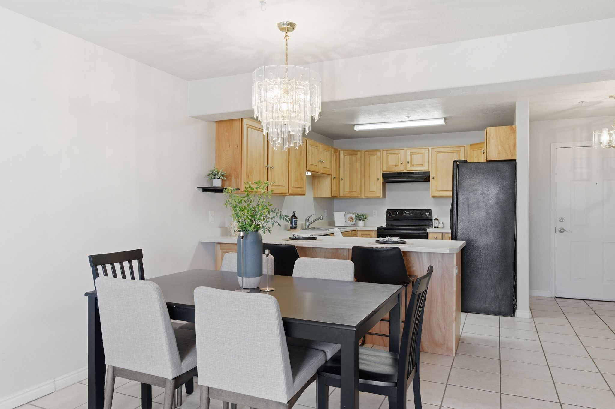 Dining area with contemporary chandelier and tile flooring, positioned with a partial wall separating the kitchen featuring wood cabinetry, a black appliance suite, and a breakfast bar