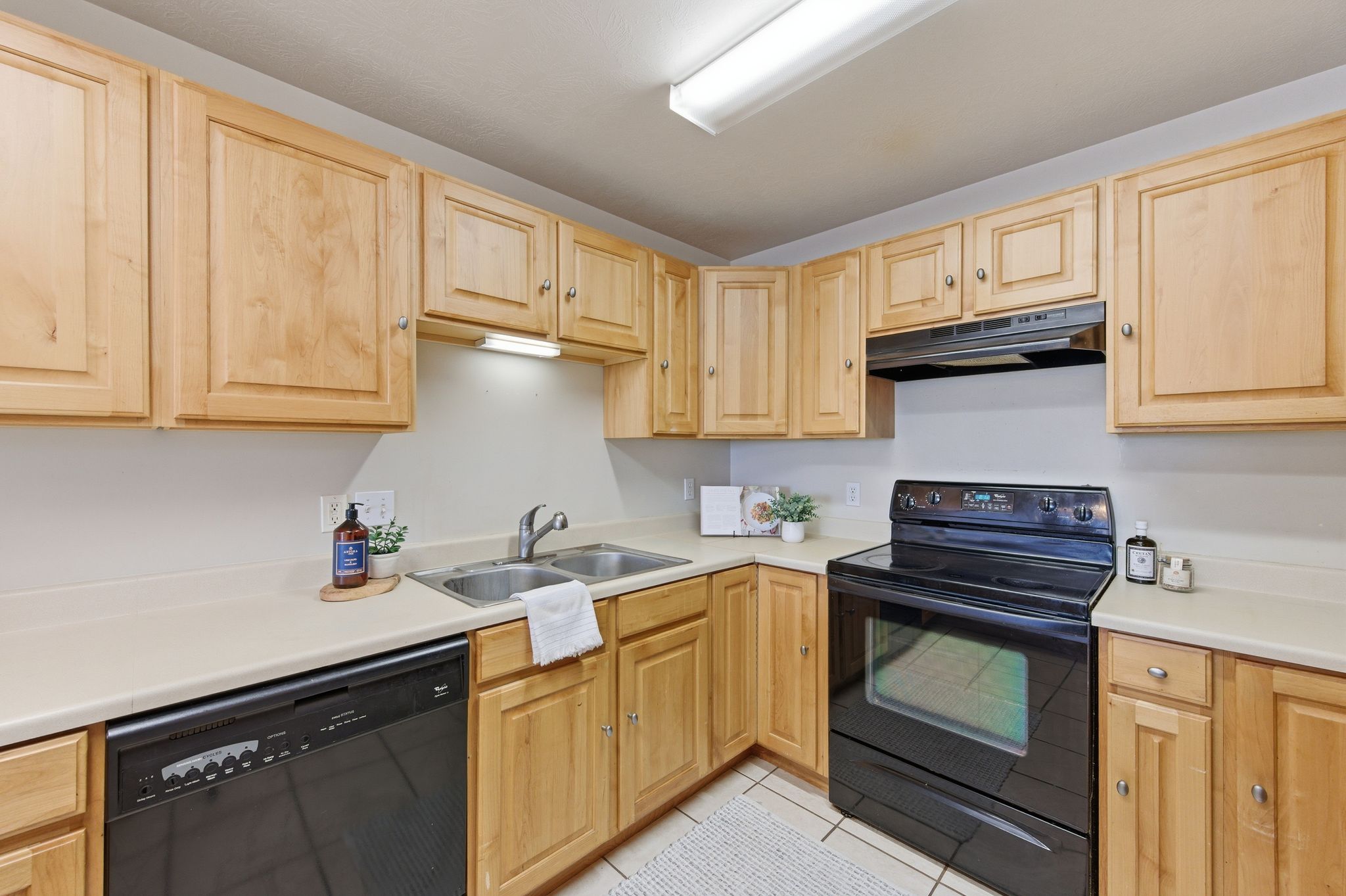 Kitchen featuring light wood cabinetry, light countertops, dual basin stainless steel sink, black electric range with overhead vent, and black dishwasher