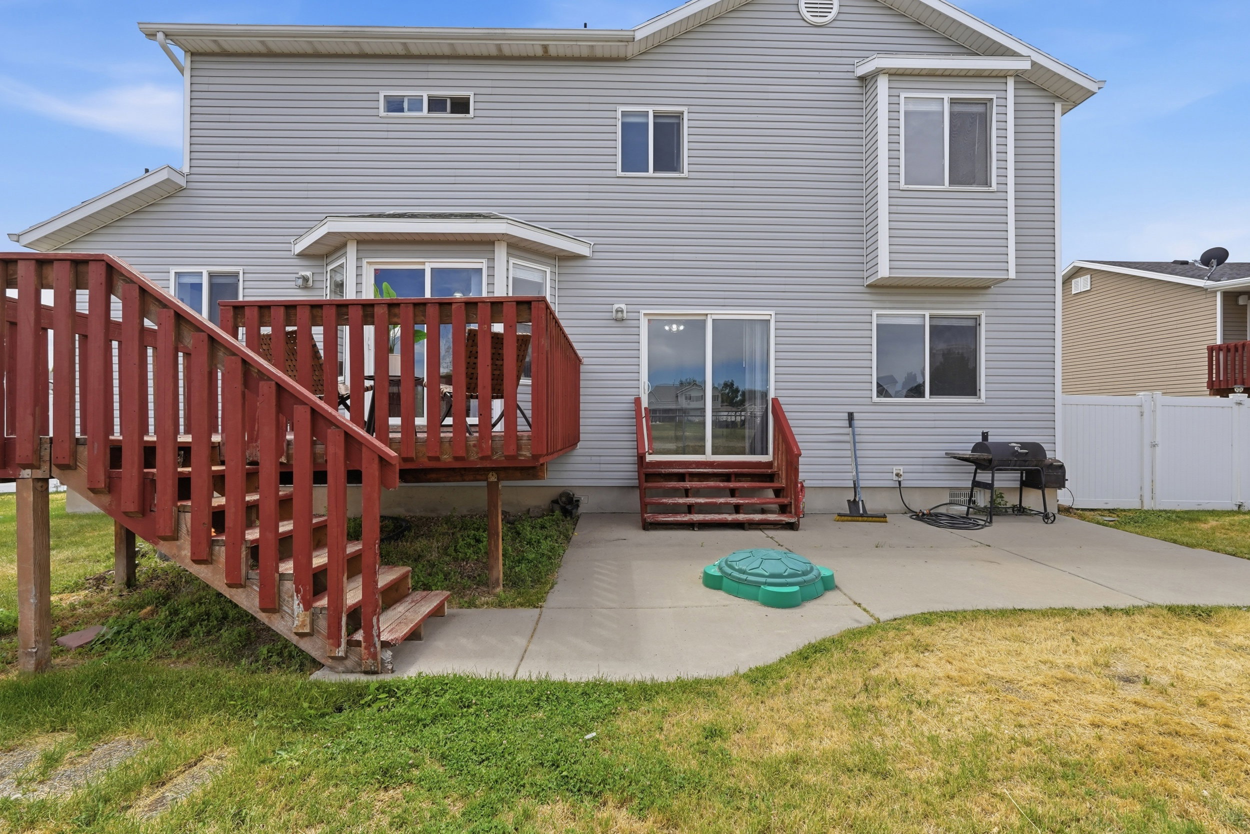Rear exterior with light gray siding, featuring a wood deck with red-stained railings and stairs, a concrete patio, and a sliding glass door
