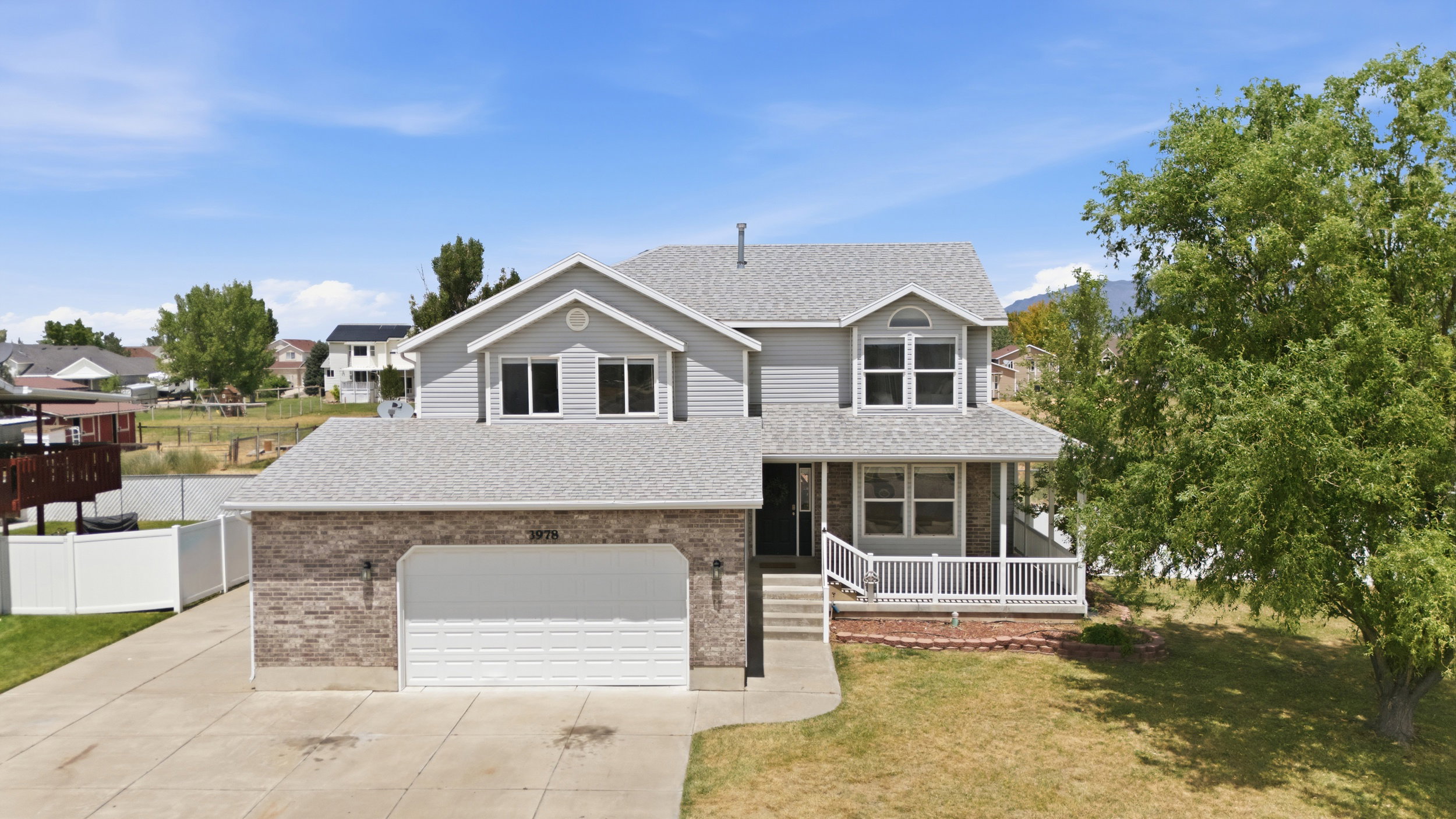 Two-story residence featuring light gray siding with brick accents, a full-width front porch with white railings, an attached two-car garage, and a concrete driveway