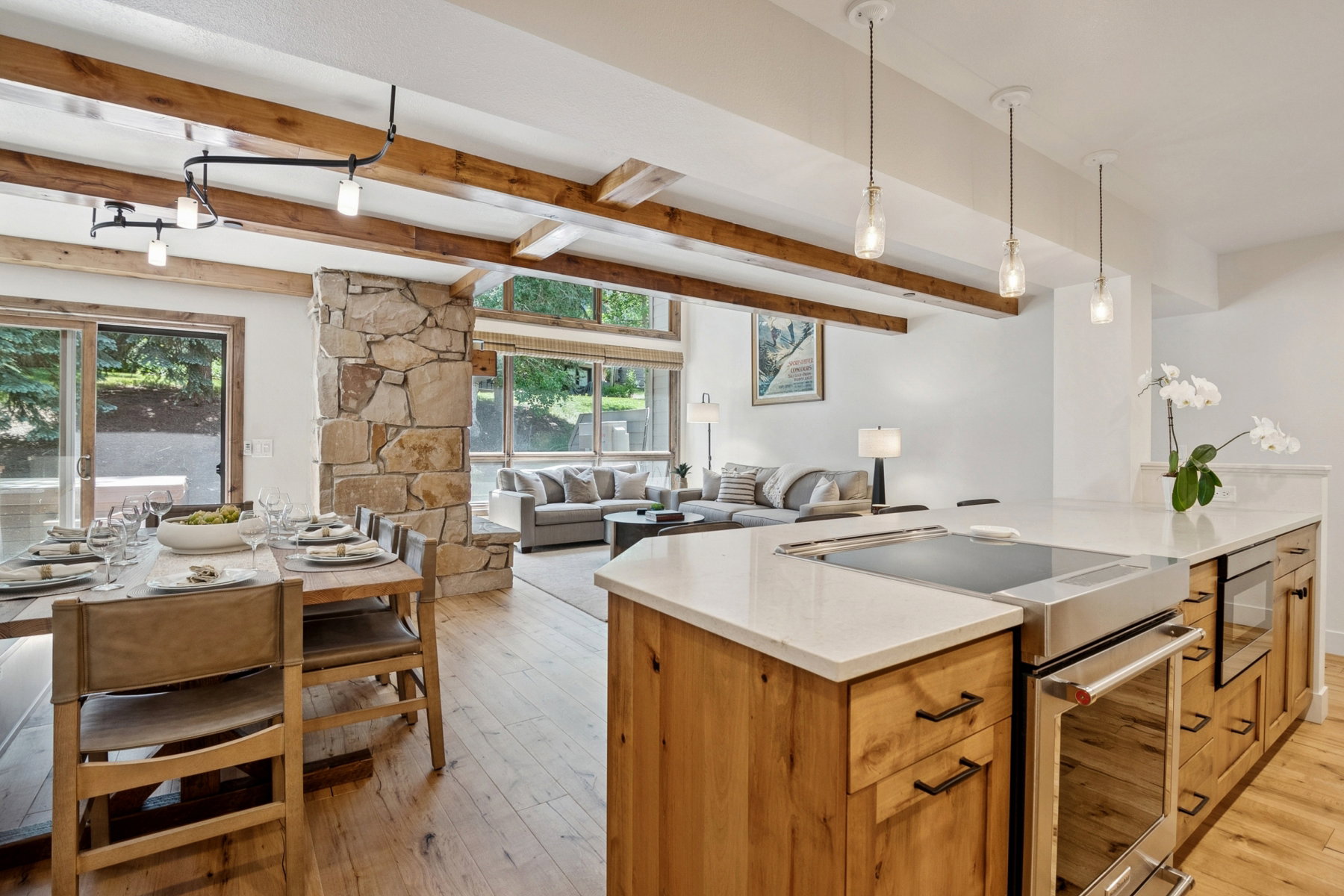 Open-concept living area with wood-finish flooring, exposed wood beams, a stone accent wall, and a kitchen island with integrated cooktop and oven