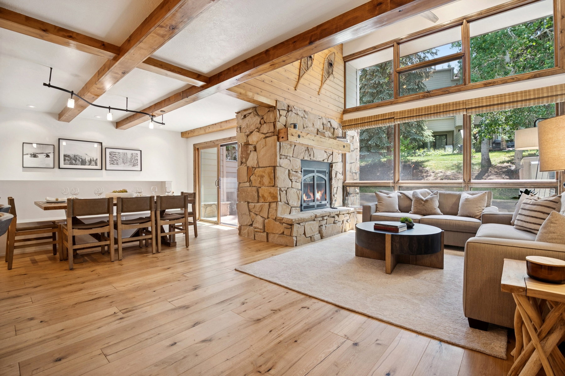 Vaulted ceilings with exposed wood beams, a prominent stone fireplace, and expansive windows characterize this living area