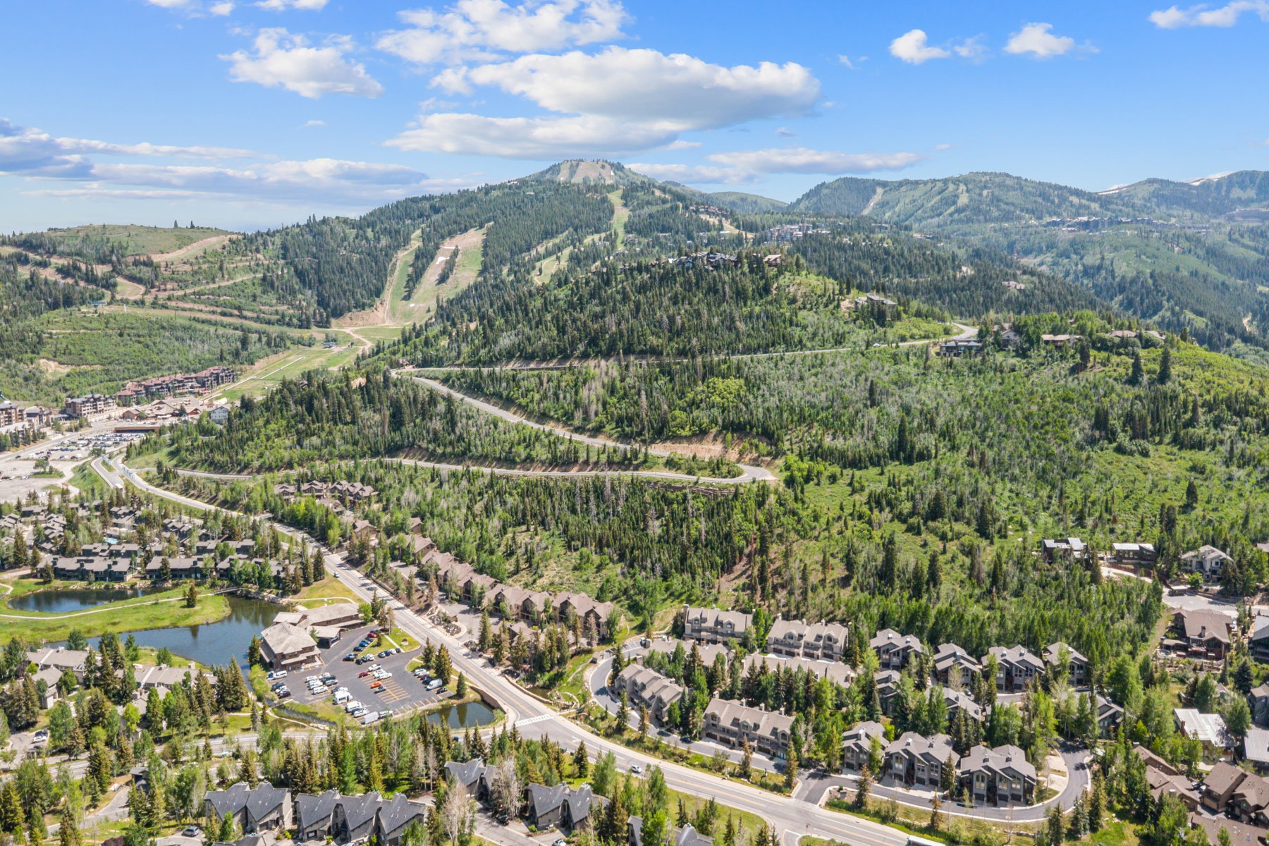 Expansive aerial showcasing a mountainous landscape with ski slopes, dense evergreen forests, and a community featuring residential properties, roads, and a pond