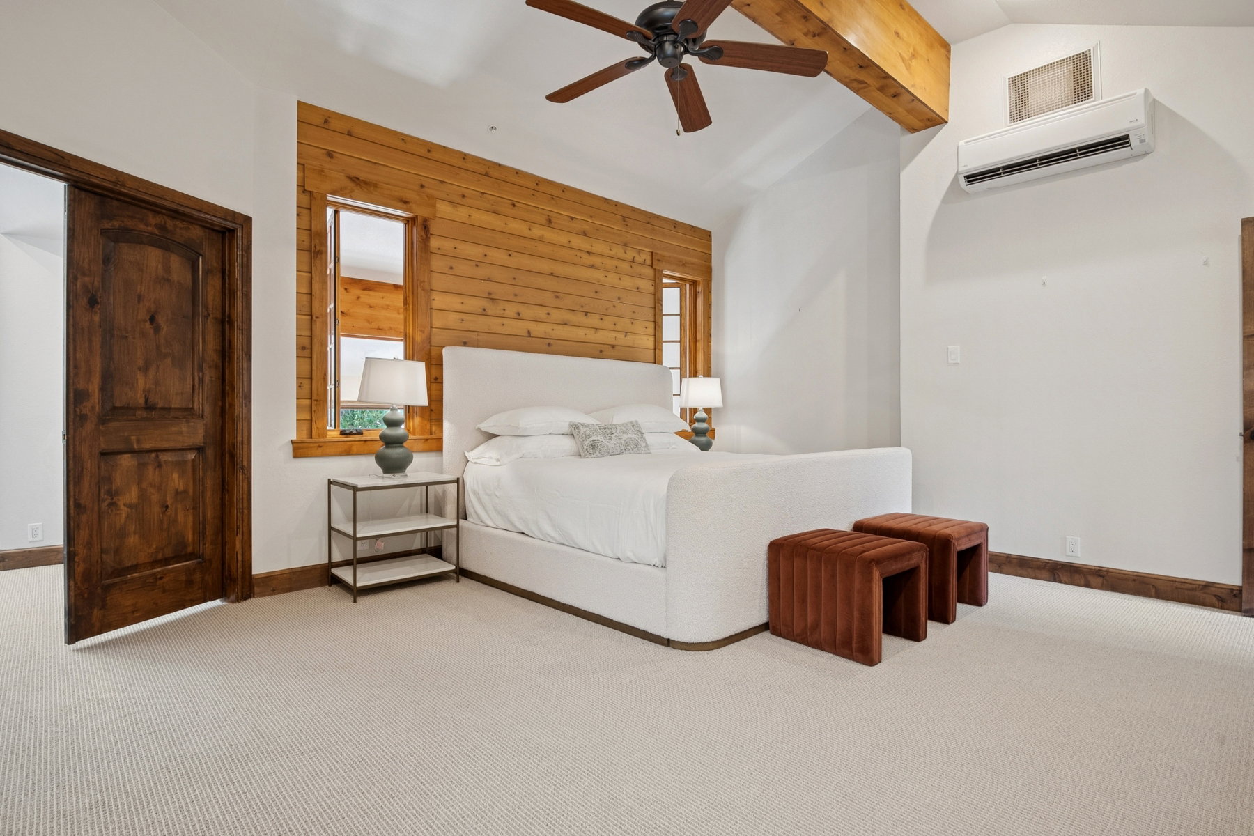Bedroom with light-toned carpeting, wood-paneled accent wall, exposed wood beam, ceiling fan, and dark wood door