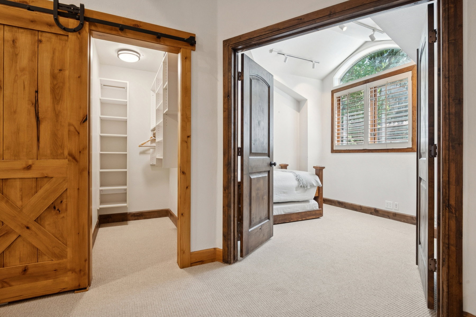 Carpeted interior featuring a rustic wood barn door, built-in closet shelving, dark wood trim, and a window with plantation shutters and an arched transom
