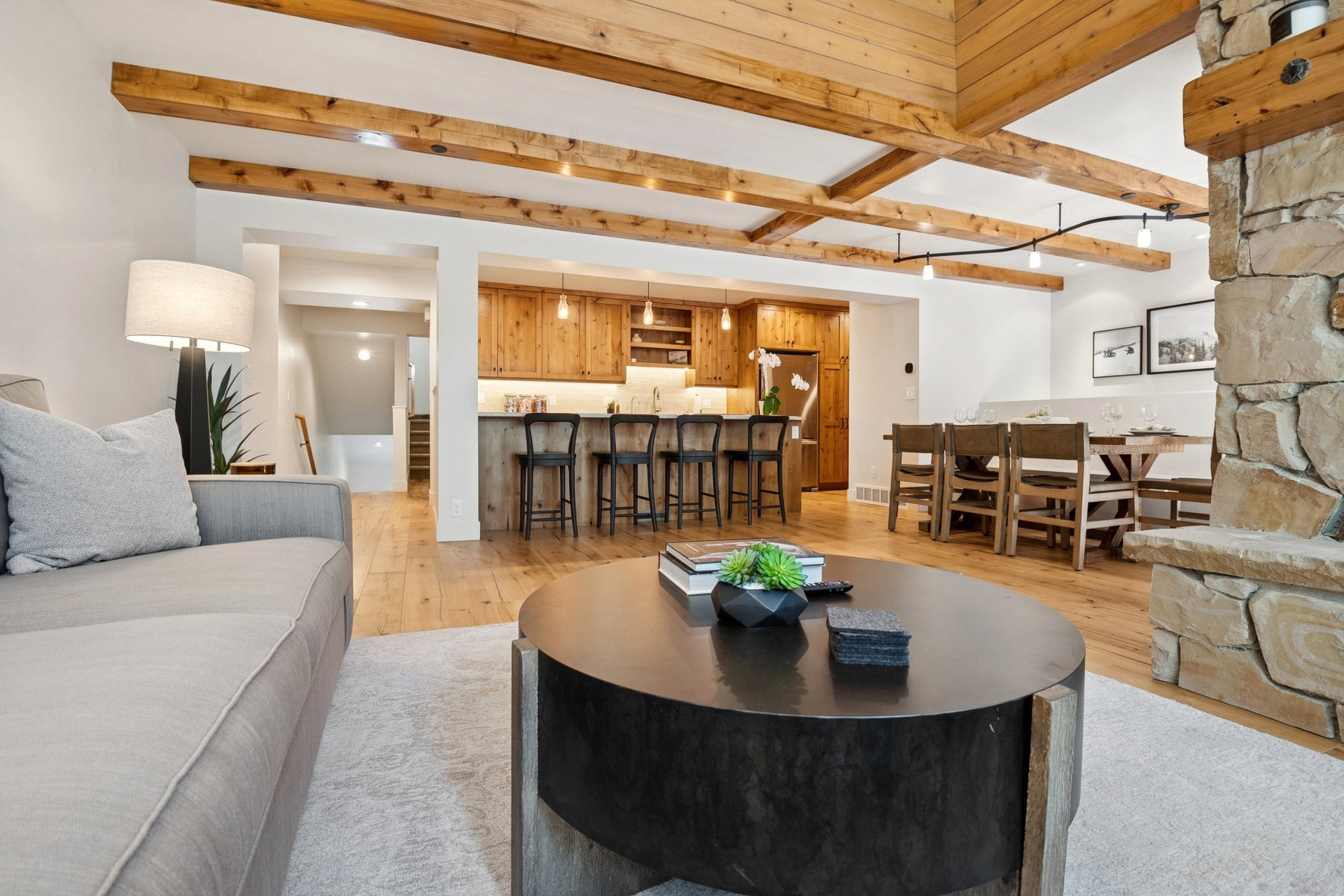 Great room featuring exposed wood ceiling beams, a stone-clad fireplace, wood-finish flooring, a kitchen with custom wood cabinetry and a peninsula with seating, and a dining area