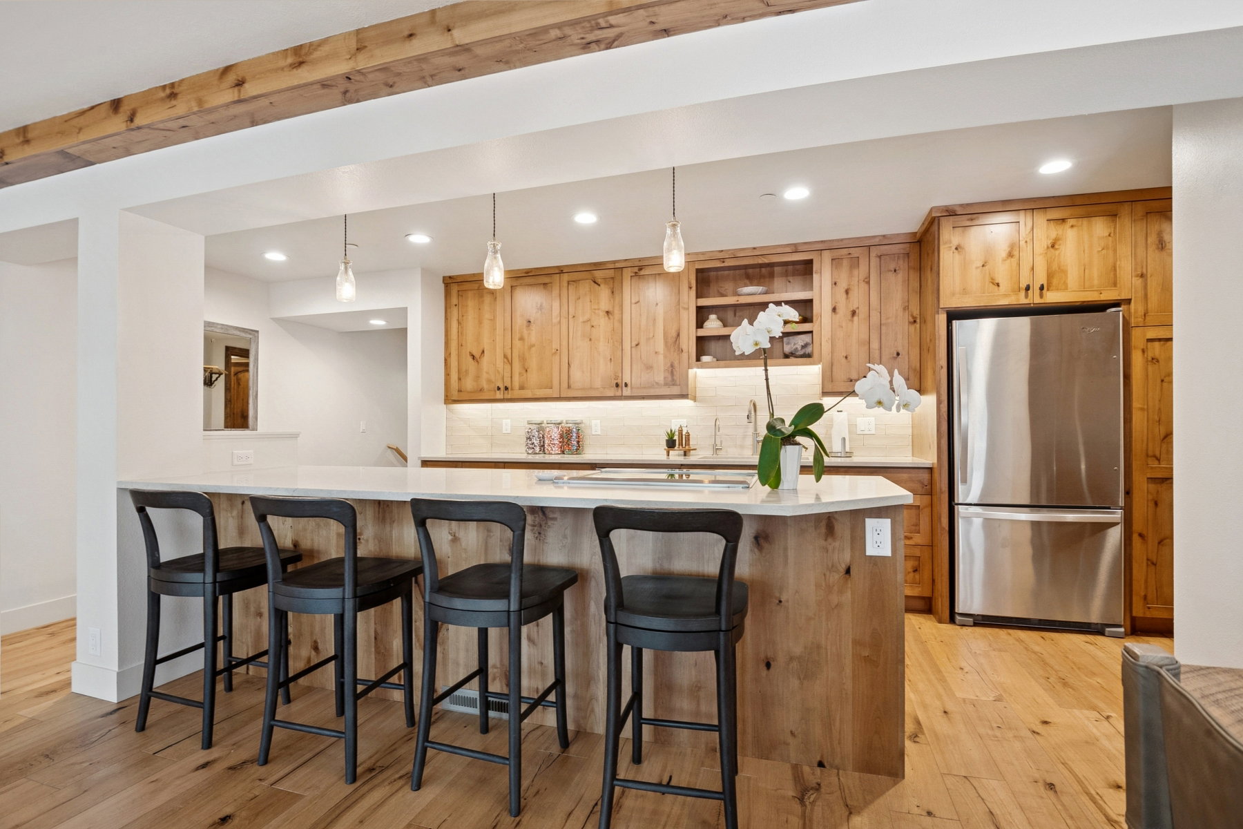 Spacious kitchen featuring an extended island with a light-toned countertop and seating for four, wood-finish flooring, exposed wood ceiling beams, natural wood cabinetry, and stainless steel appliances