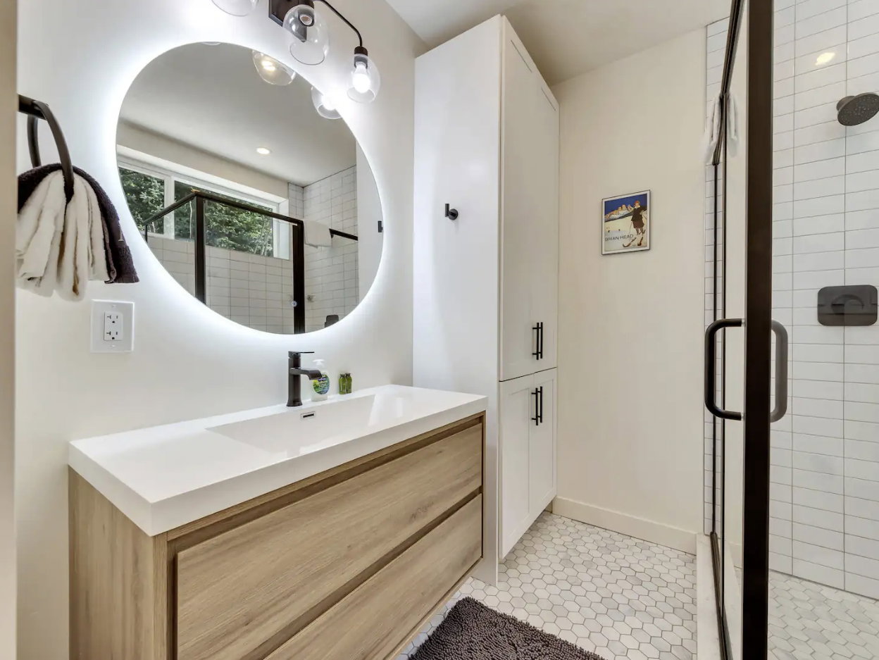 Contemporary bathroom featuring a light wood-finish vanity with a white countertop and a vessel sink, complemented by a backlit oval mirror