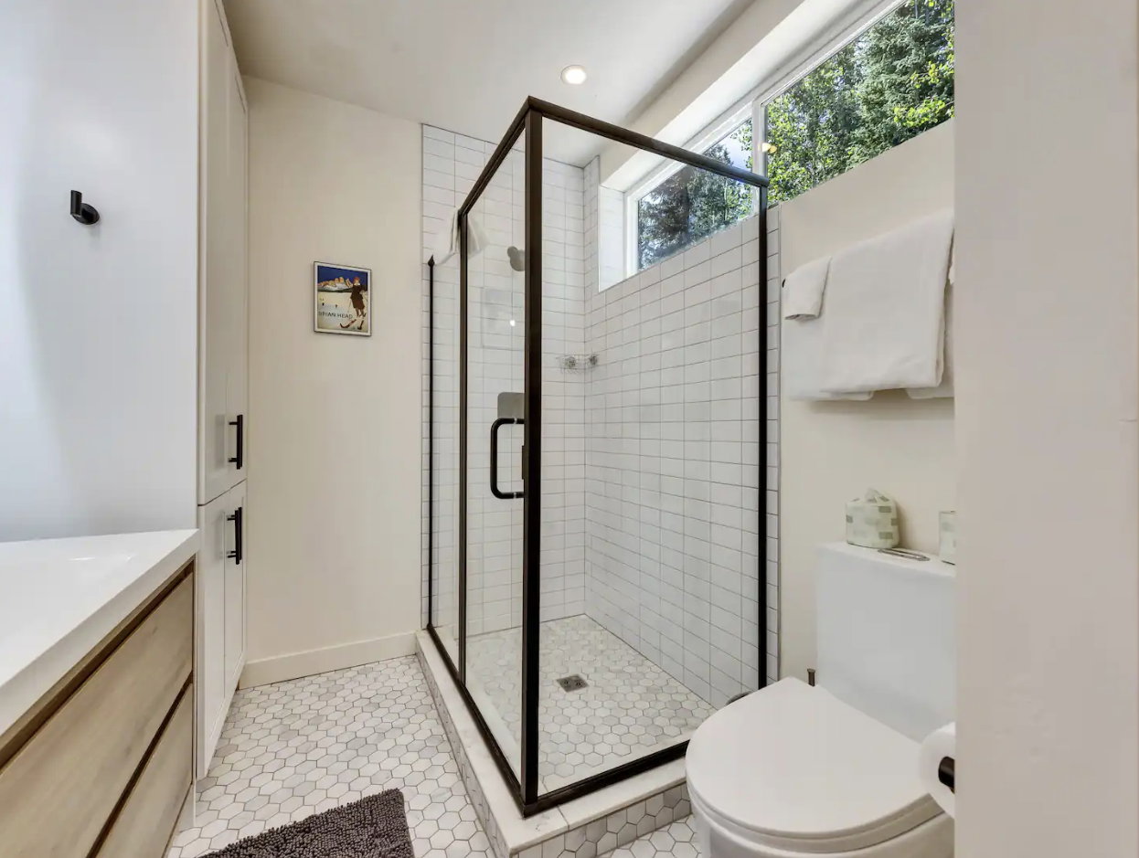 Modern bathroom featuring a glass-enclosed shower with subway tile surround, white hexagonal floor tiling, a single vanity with wood-finish cabinetry and white countertop, and a white toilet